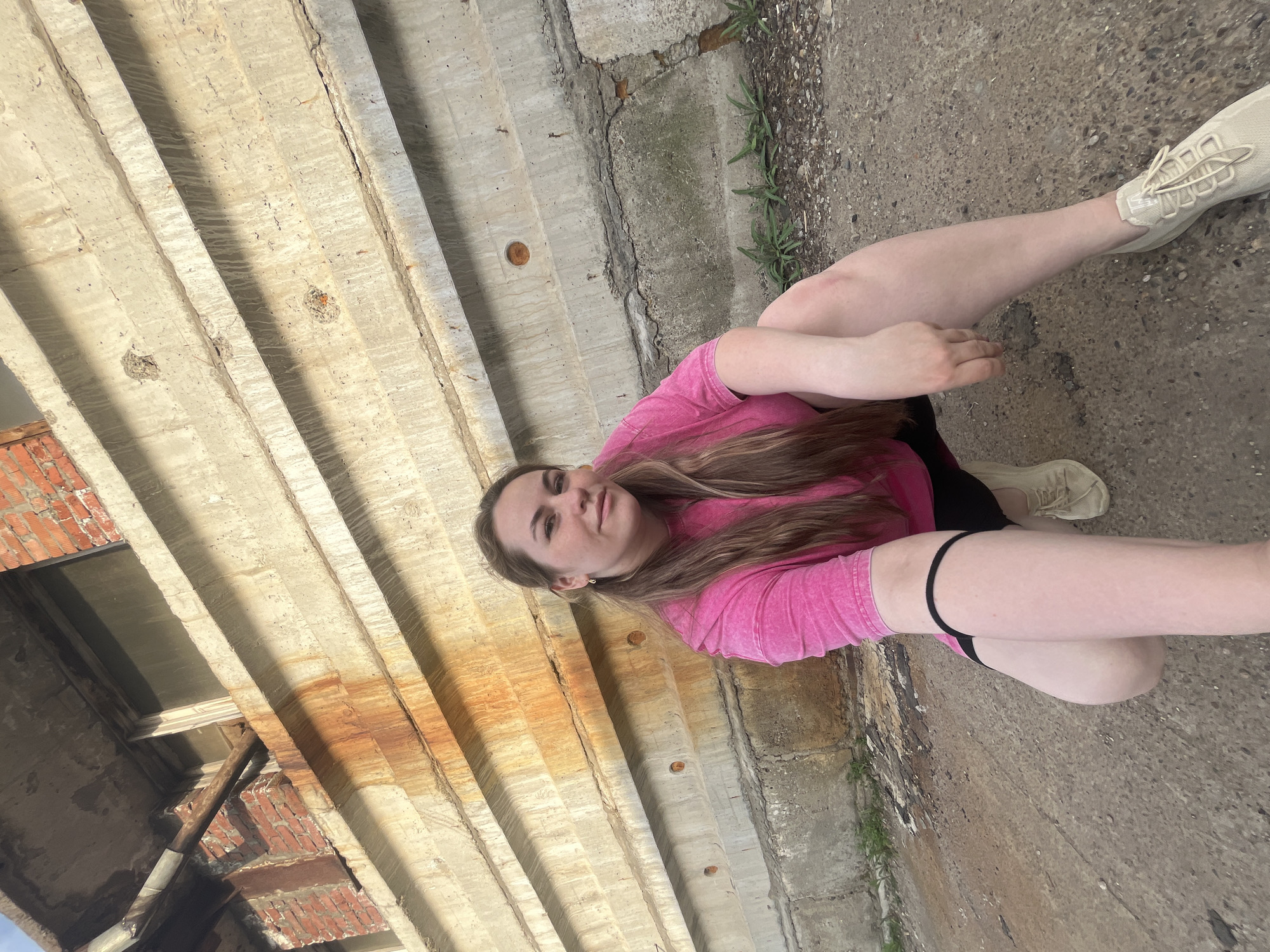 A young woman is sitting on a concrete surface in Chaikovsky, Russia. The woman is wearing a pink shirt, black shorts, and light-colored sneakers. She has long brown hair and is facing the camera. A concrete structure with rusty stains is behind her, and greenery is visible on the right side of the frame. The ground appears to be made of concrete.
