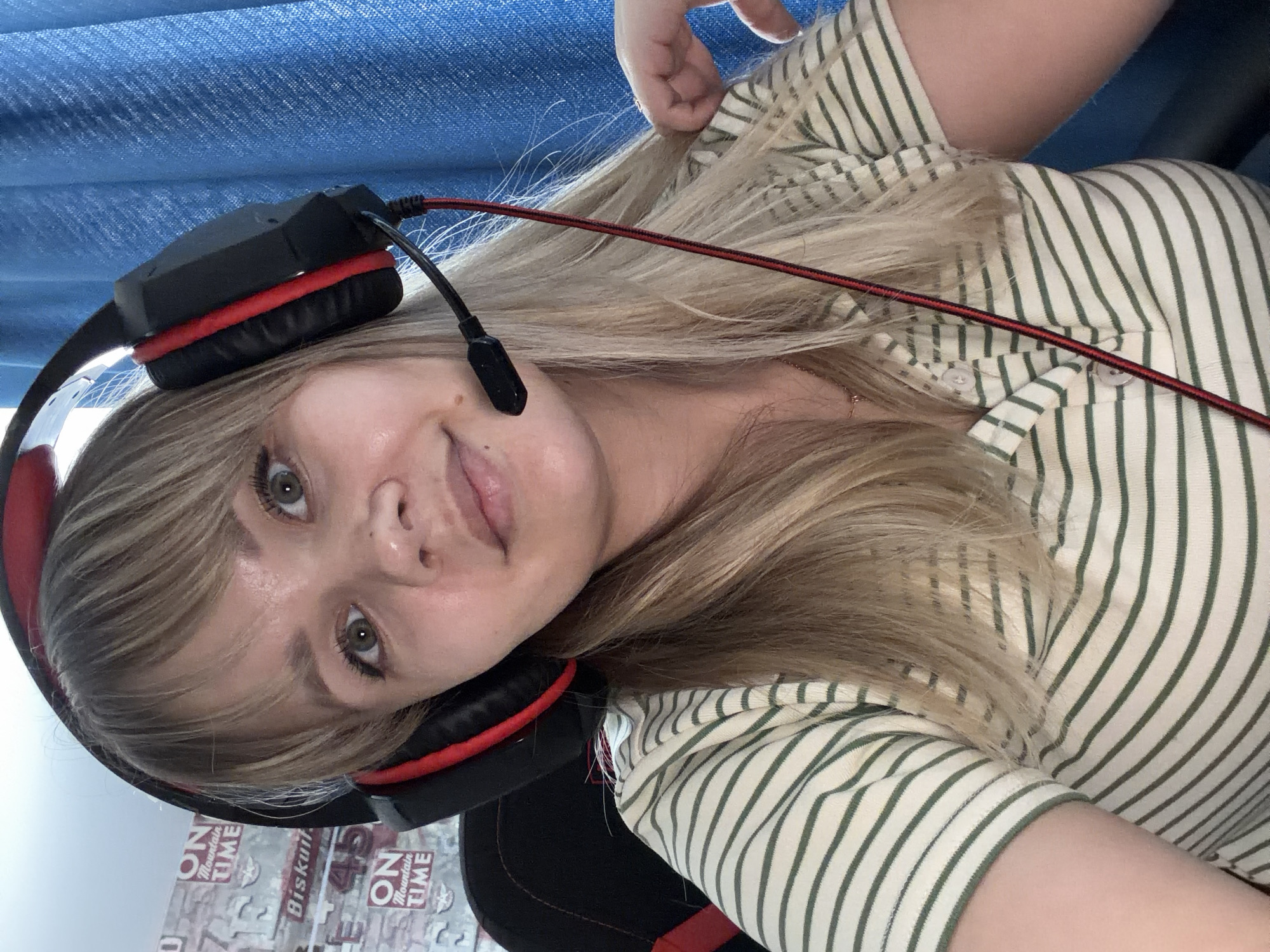 An image, rotated clockwise, depicts a young woman from the chest up, wearing large gaming headphones with a microphone. She has fair skin, light blonde hair with bangs, and light-colored eyes. Her expression is a soft, closed-mouth smile, and she looks directly towards the camera. She is wearing a cream or off-white long-sleeved shirt with thin, dark green vertical stripes and visible buttons near the collar. Her left hand is subtly touching her hair near the top right of the frame.
The headphones are black with bright red accents on the earcups and a red braided wire extending from the left earcup. A black microphone boom extends from the left earcup towards her mouth.
The background primarily consists of a textured blue fabric, possibly a curtain or blind, at the top. Below the person, there's a wall or surface adorned with text and graphics, including repetitive phrases like "ON MOUNTAIN TIME" and the word "Biskuit" along with the numbers "43". A portion of a black and red chair back is also visible beneath her. The scene appears to be indoors, likely a casual setting such as a home or personal workspace, suggesting the woman might be engaged in gaming, streaming, or an online conversation.