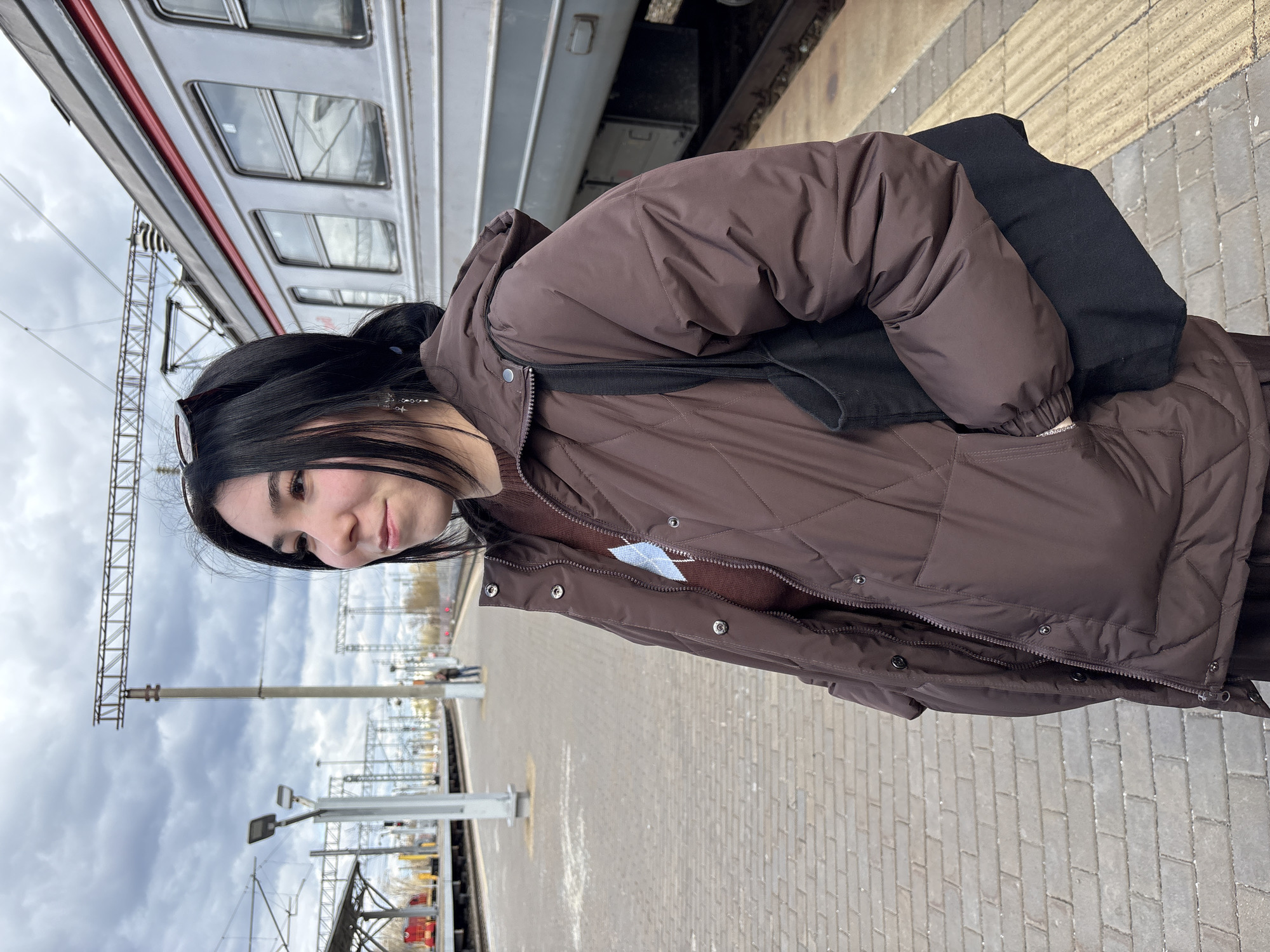 A young woman with dark hair stands on a platform next to a train. She wears a brown quilted jacket and a black bag is slung over her shoulder. The platform is made of gray rectangular stones. The train car is silver with a red stripe. The background shows an overcast sky and train tracks with overhead electrical wires. This photo was taken at the Kazan, Russia railway station.