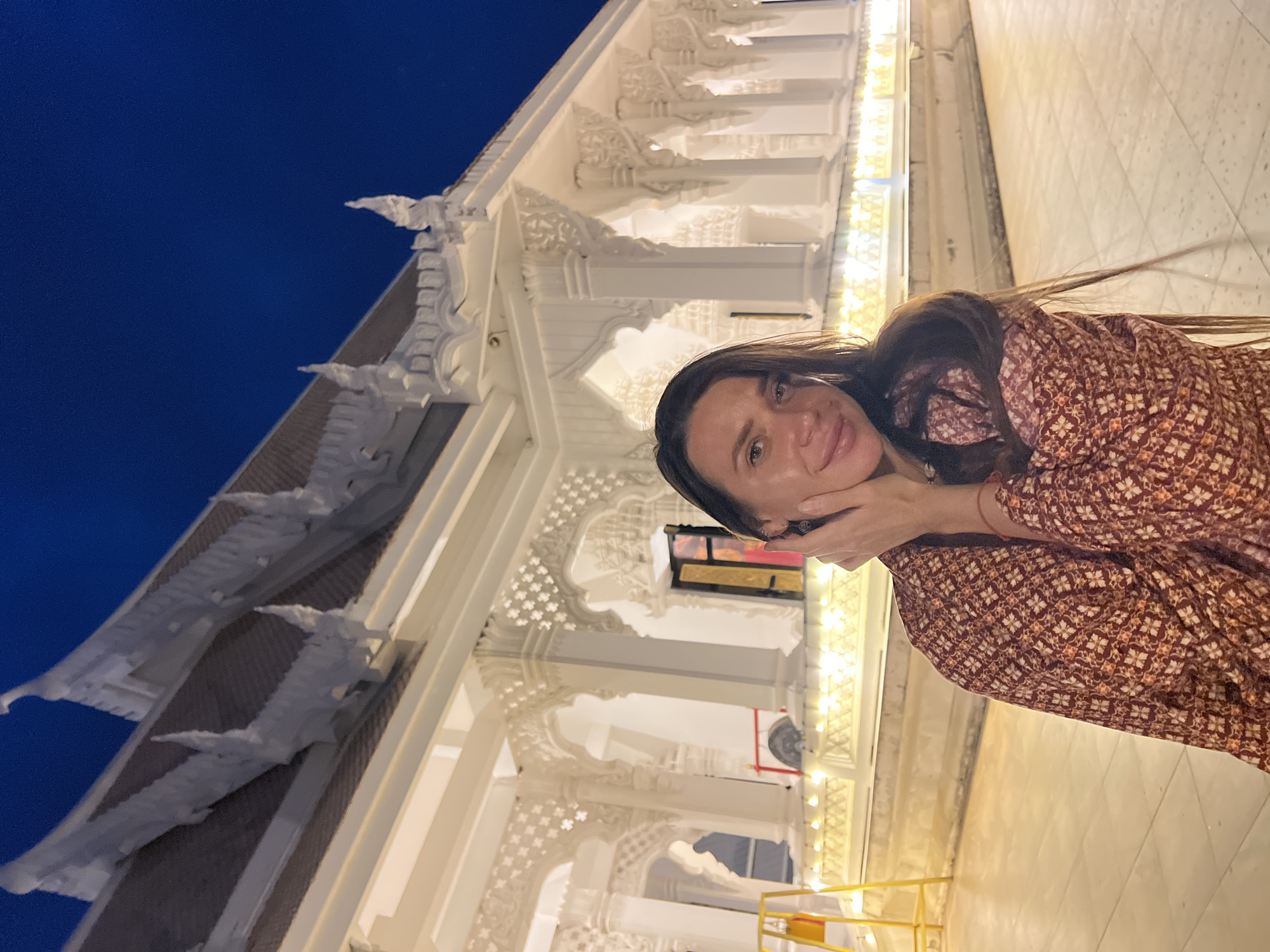 A young woman is posing in front of a building in Baan Bo Phut, Thailand. She is holding her hands to her face and smiling at the camera. The building behind her is intricately decorated with white carvings and illuminated with lights. The sky is a dark blue in the upper left corner. The woman is wearing a long-sleeved patterned dress. The ground is tiled in a light color. A yellow pole is visible in the lower right corner of the image.