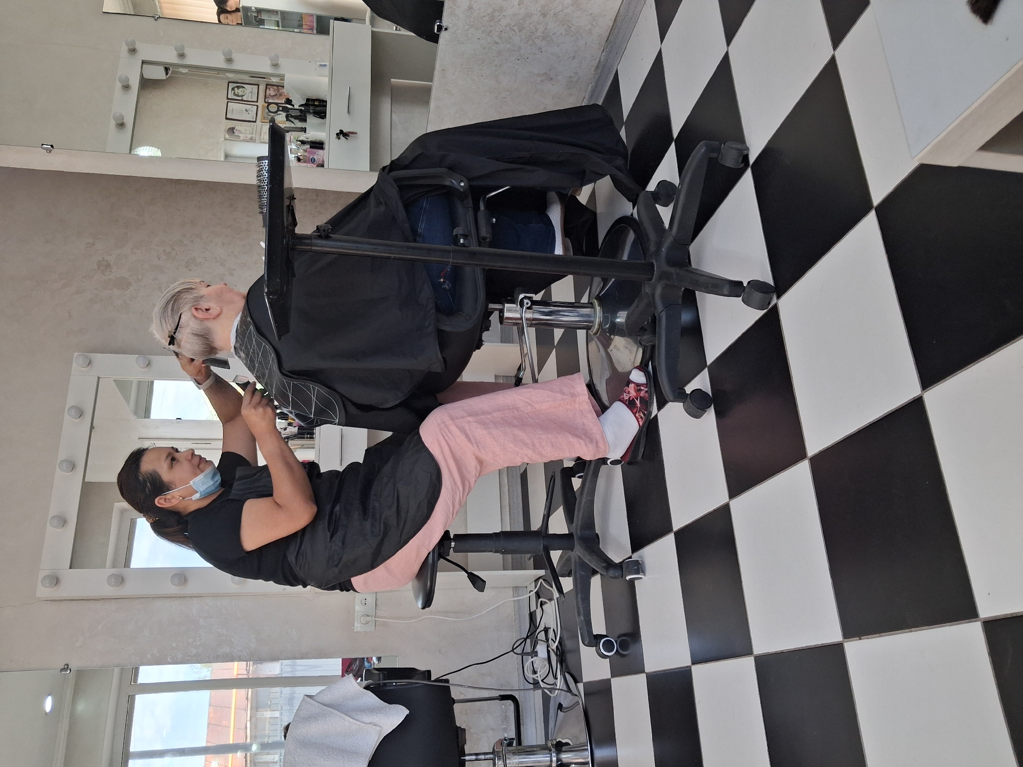 A woman with her hair in a ponytail, wearing a black t-shirt, a black apron, pink pants, and floral-patterned slip-on shoes, is seated on a black rolling stool. She is masked and using scissors and a comb to cut the hair of another person, who is wearing a black cape. This person is seated in a chair and has short, blonde hair. The scene takes place inside a brightly lit hair salon. The floor is tiled with a black and white checkerboard pattern. In the background, there are mirrors with lights around them, shelves with various hair products, and windows that look out onto a building and some trees. The setting appears to be a commercial establishment within the city of Tuzdybastau, Kazakhstan.