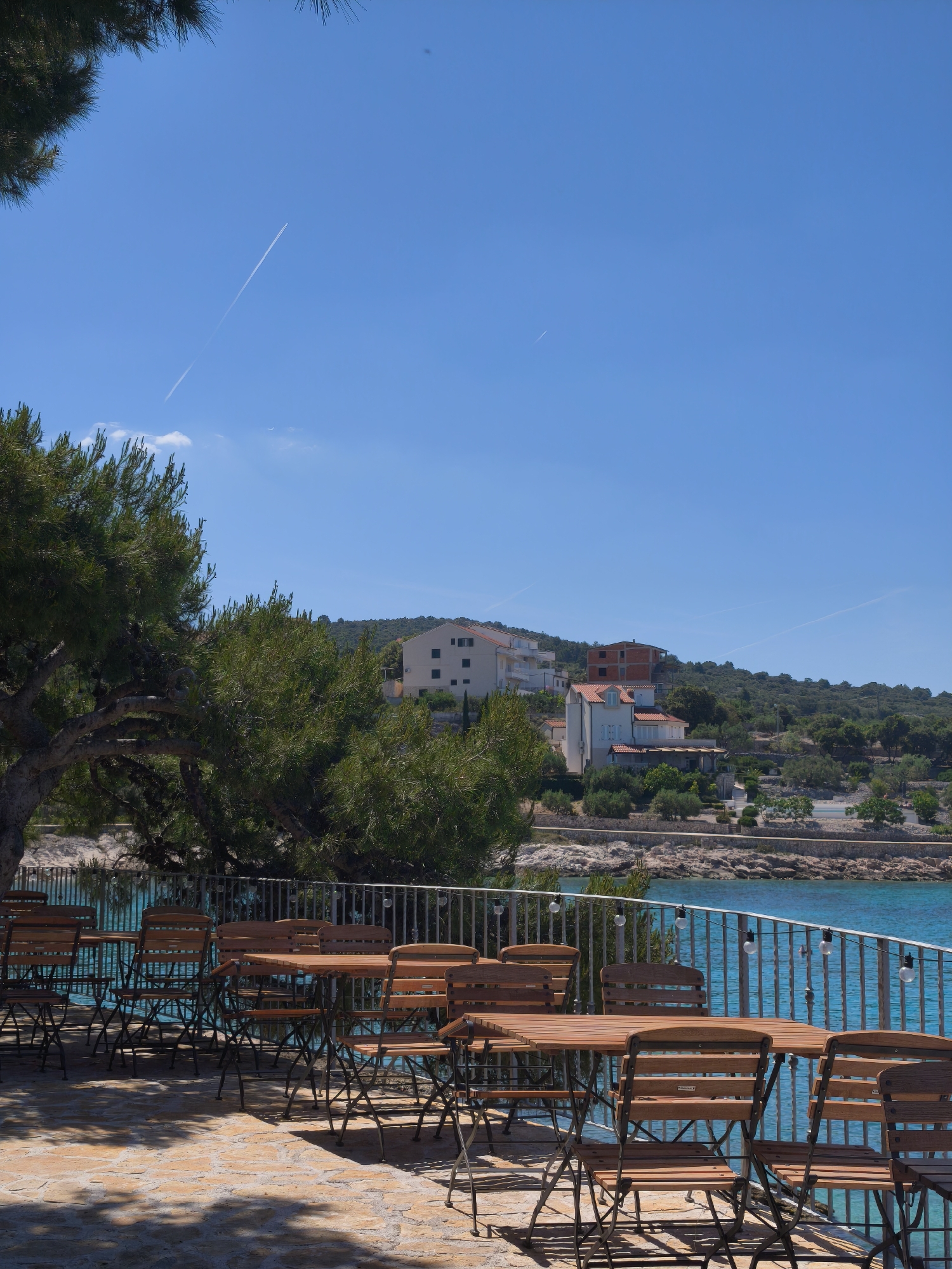 The image depicts an outdoor cafe or restaurant setting on a sunny day, likely situated in an area with coastal views. The scene features multiple wooden tables and chairs arranged on a stone terrace. A metal railing borders the terrace, offering a view of the turquoise water and the shoreline in the background. Several buildings, including houses with red-tiled roofs and a building under construction, are visible on the hillside across the water. Vegetation, including trees and shrubs, is interspersed among the buildings. A tree on the left side of the image partially obscures the view, casting shadows on the terrace. In the clear blue sky, contrails from aircraft can be seen. The location is in the city of Unknown, Unknown.