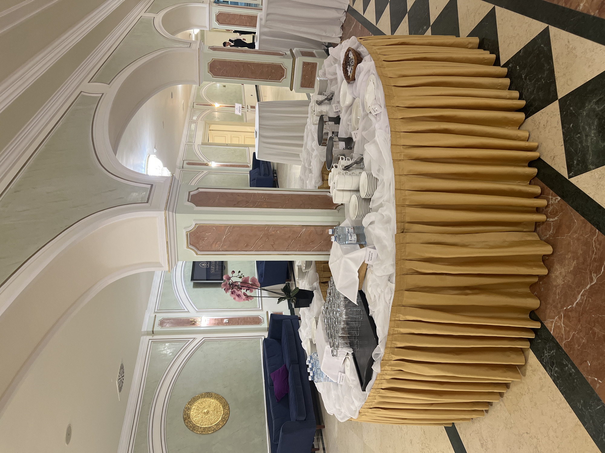 This image depicts a catering setup in what appears to be a banquet hall or event space, possibly at the Saryarqay Event Hall in Astana, Kazakhstan, given the visible branding. The scene is centered around a large, round table draped with a golden, pleated skirt and a white tablecloth. On the table are arrangements of glassware, stacks of plates and cups, serving dishes, bottles of water, and a small basket of sugar packets. In the background, several tall, round tables covered with white cloths are visible, along with a dark blue sofa with a purple cushion against a wall decorated with ornate green and gold paneling. Archways and columns contribute to an elegant interior design. One corner of the room shows a television screen displaying the "SARYARQAY EVENT HALL" logo and the words "EVENT HALL." A person in a black vest and white shirt is partially visible in the background. The flooring is a patterned mosaic of marble tiles in shades of cream, dark green, and red.