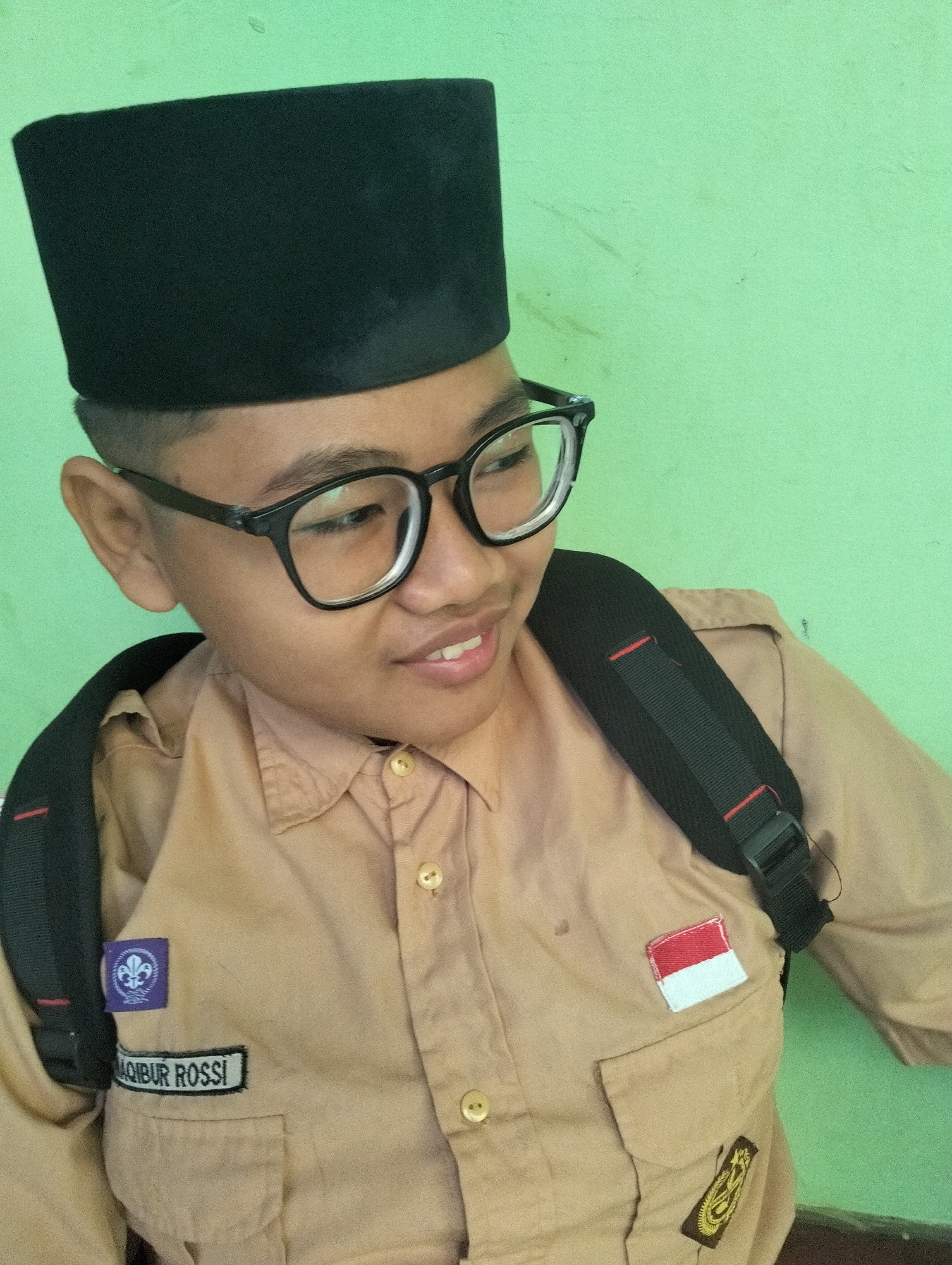 A medium shot shows a young Indonesian boy, likely a student, wearing a tan uniform and a black peci hat. He is also wearing black-rimmed glasses and a backpack. On his left uniform breast is a purple patch with a fleur-de-lis symbol and a name tag reading "MOIBUR ROSSI". To the right of that is a small Indonesian flag patch. The background is a plain, light green wall. The image appears to have been taken indoors or in a sheltered outdoor area. This scene is consistent with a school or scouting activity. The boy is looking to his right and smiling.