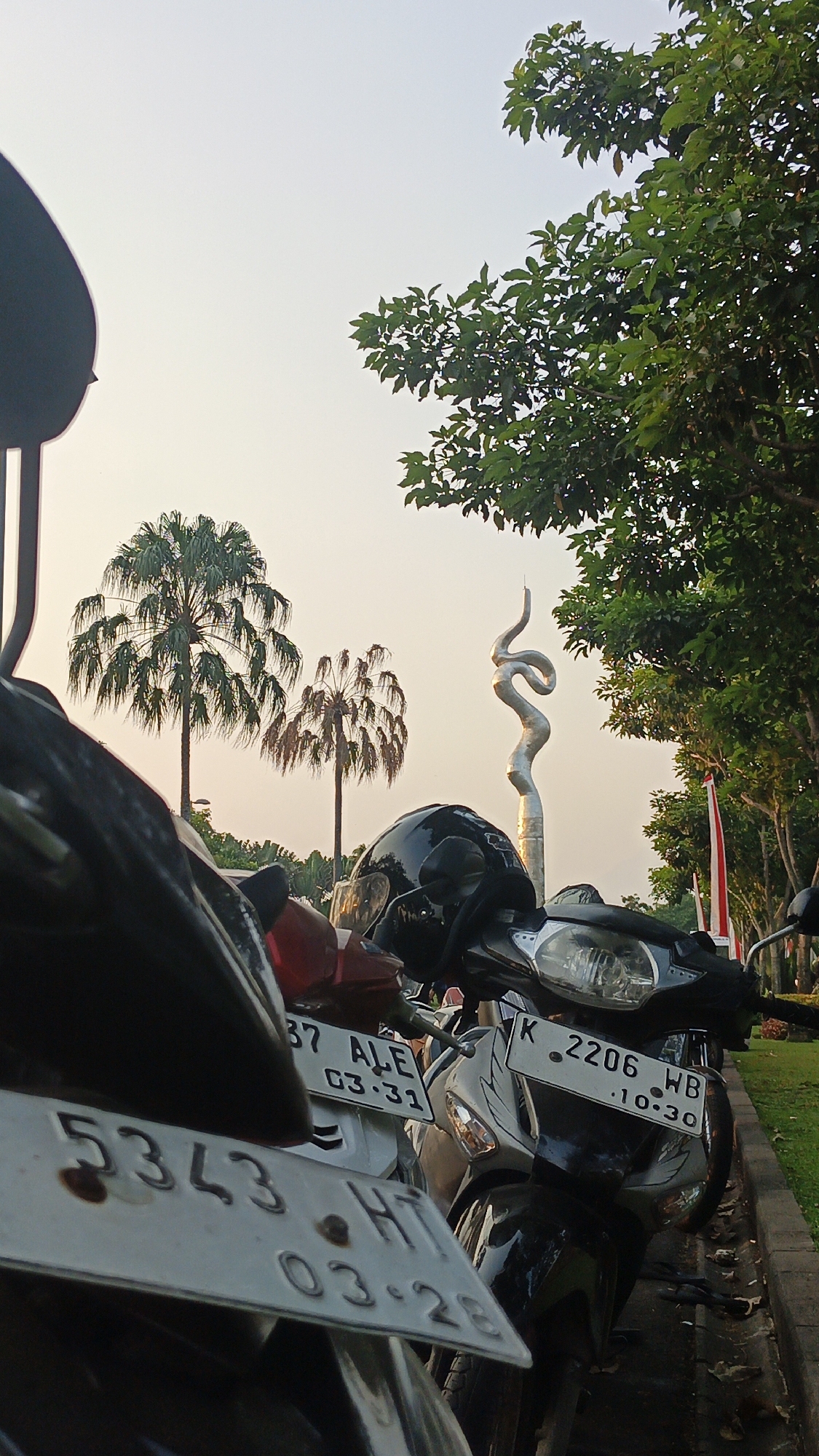The image depicts a row of parked motorcycles in front of a park-like area. In the background, there is a distinctive silver sculpture that appears to be an abstract depiction of a snake or intertwined ribbons, with two palm trees to its left. The sky is a pale yellow, suggesting either dawn or dusk. The foreground is dominated by the front of the motorcycles, with their license plates visible. One license plate reads "5343 HT 03-28". Another plate visible on a motorcycle further back says "37 ALE 03-31". A third plate reads "K 2206 WB .10.30". A black helmet rests on the handlebars of one of the motorcycles. Green trees and some red and white flags are visible on the right side of the image, lining a path or road. The overall scene appears to be a public space, possibly a parking area near a monument or recreational spot within Bacin, Indonesia, given the context of the sculpture and tropical vegetation. There are no people visible in the image.