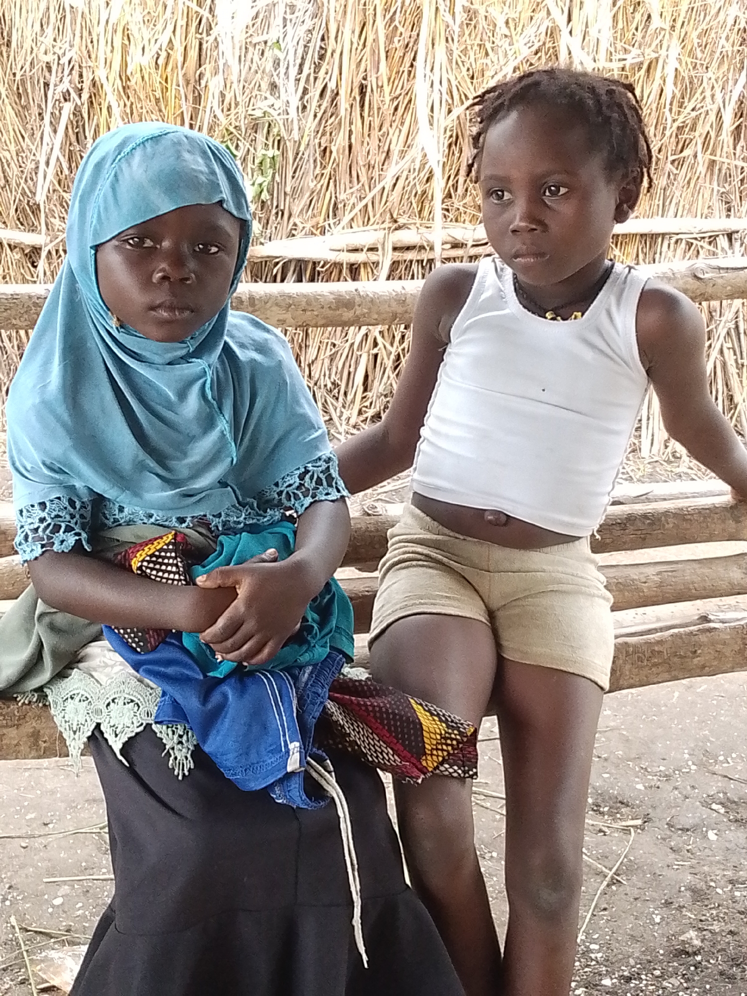 Two young Black girls are sitting on a wooden beam in front of a thatched wall. The girl on the left is wearing a light blue hijab and a dark dress, and she is holding a bundle of colorful clothes. The girl on the right is wearing a white tank top and light brown shorts, and she has a beaded necklace on. Both girls are looking away from the camera. The scene appears to be outdoors, in a rural setting, possibly in Otulu, Nigeria, judging by the thatched wall and the dry, dusty ground.