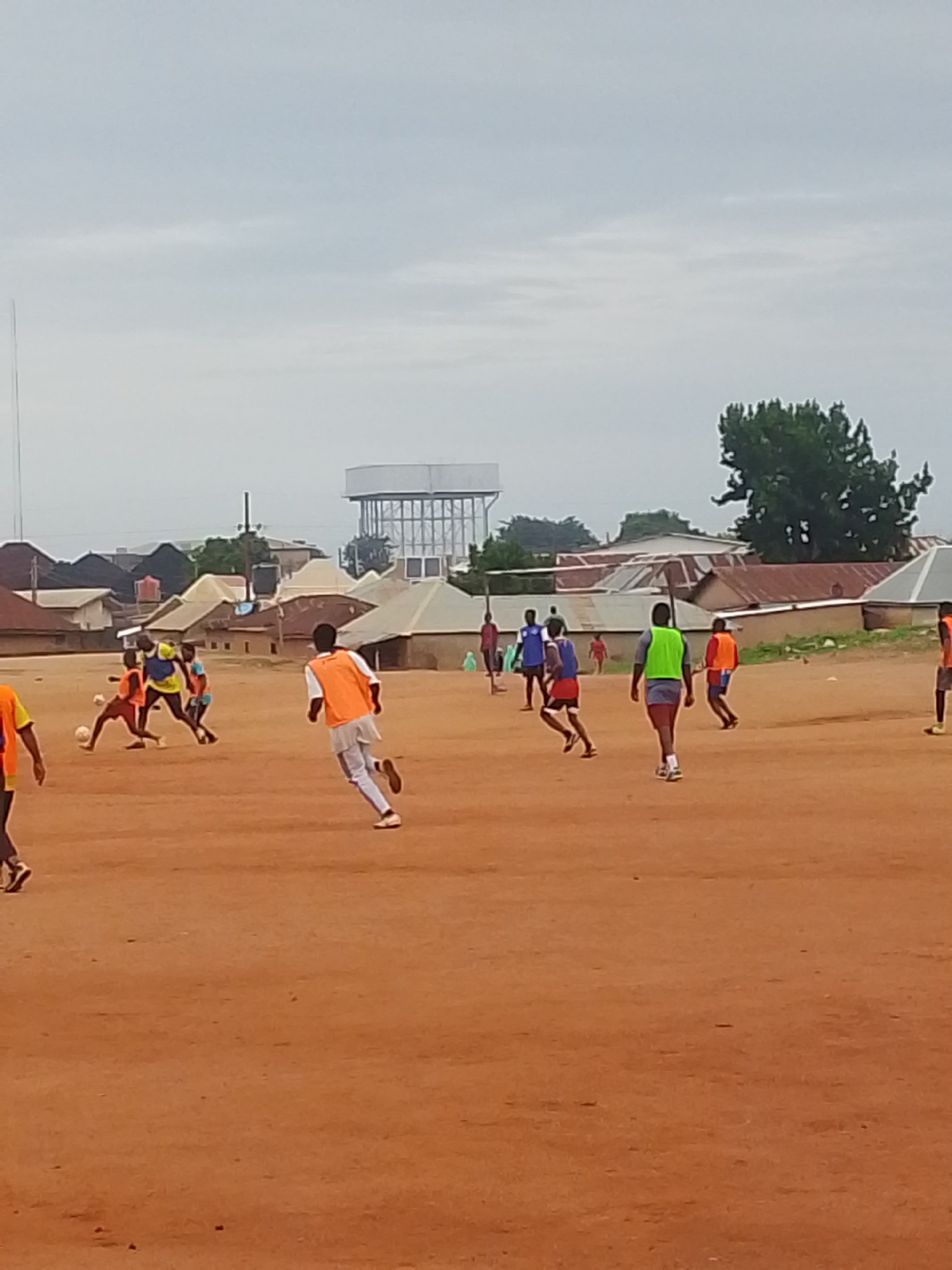 The image depicts a soccer game taking place on a dirt field in Bukuru, Nigeria. Several players wearing different colored jerseys are actively playing, with some running and others interacting with the soccer ball. In the background, there are buildings with tin roofs, a water tower, and trees, typical of the urban environment. The sky is overcast.