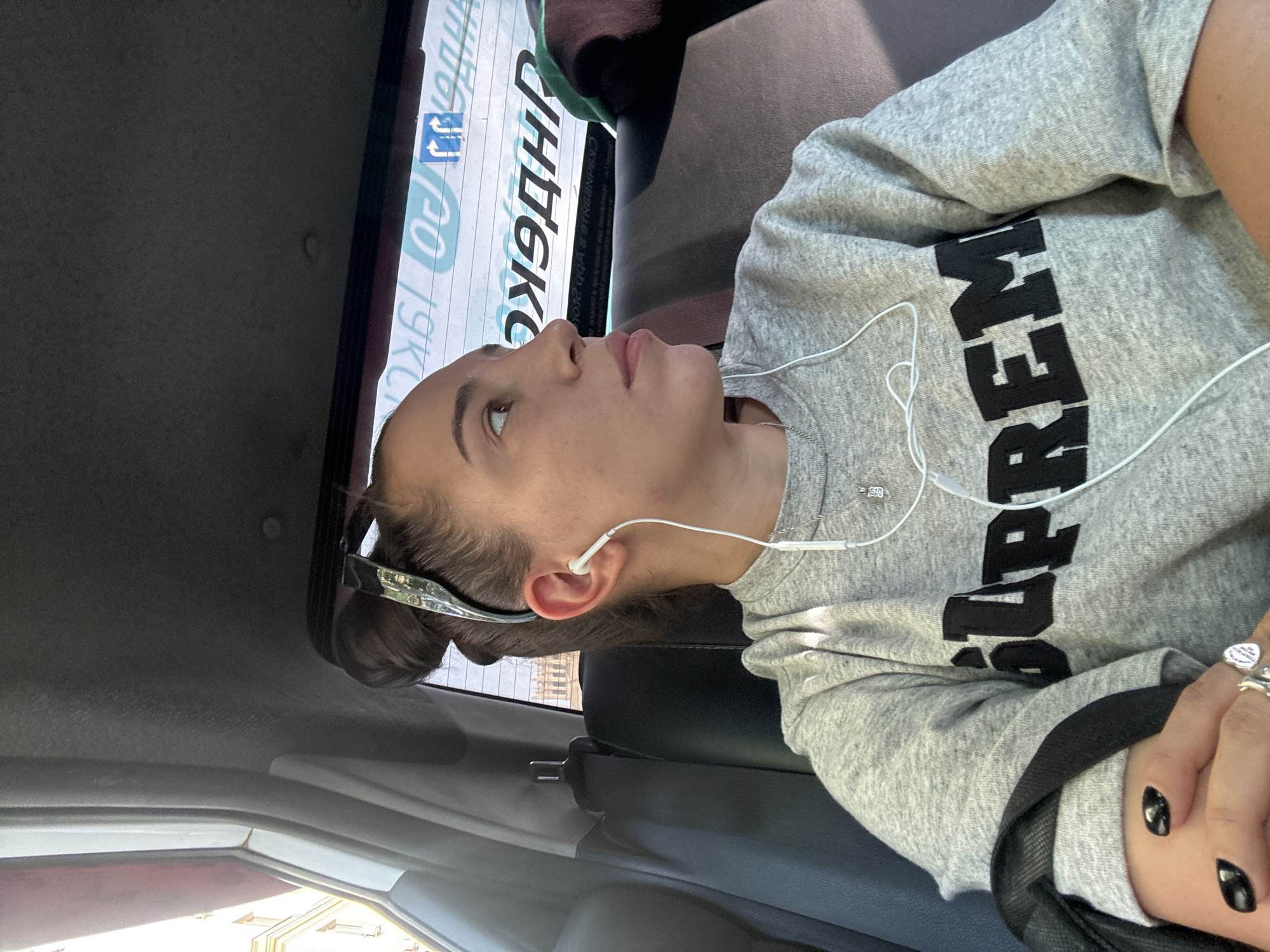 A woman with brown hair pulled back in a bun is seated in the back seat of a car, likely in Moscow, Russia. She is wearing a grey "Supreme" sweatshirt, has headphones in her ears, and is wearing rings on her fingers. She appears to be looking out the window, which has a sign with Russian text. The car's interior is visible, including the seat and dashboard. The woman is also wearing a seatbelt.