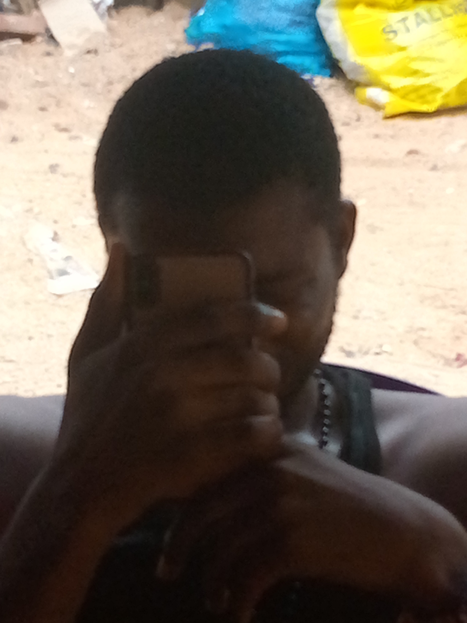 A person with dark skin and short hair is holding a phone in front of their face. The person is wearing a dark sleeveless shirt. A beaded necklace is visible around their neck. In the background, there's a yellow bag with the partial word "STALL" visible and a blue bag to its left. The location appears to be outdoors, likely in Onitsha, Nigeria.