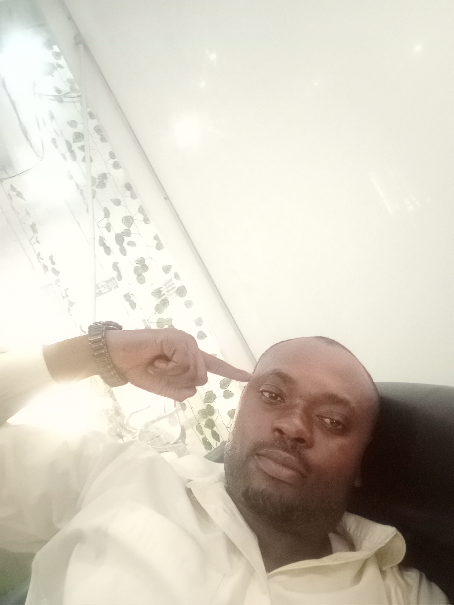 A selfie of a dark-skinned man with a shaved head, wearing a light-colored shirt and a metal watch, is taken at an upward angle. The man is pointing to the side of his head with his right index finger. Behind him are partially visible white curtains with a leaf pattern, a white wall, and a dark-colored chair. It is impossible to determine if the location is Gwarinpa, Nigeria.