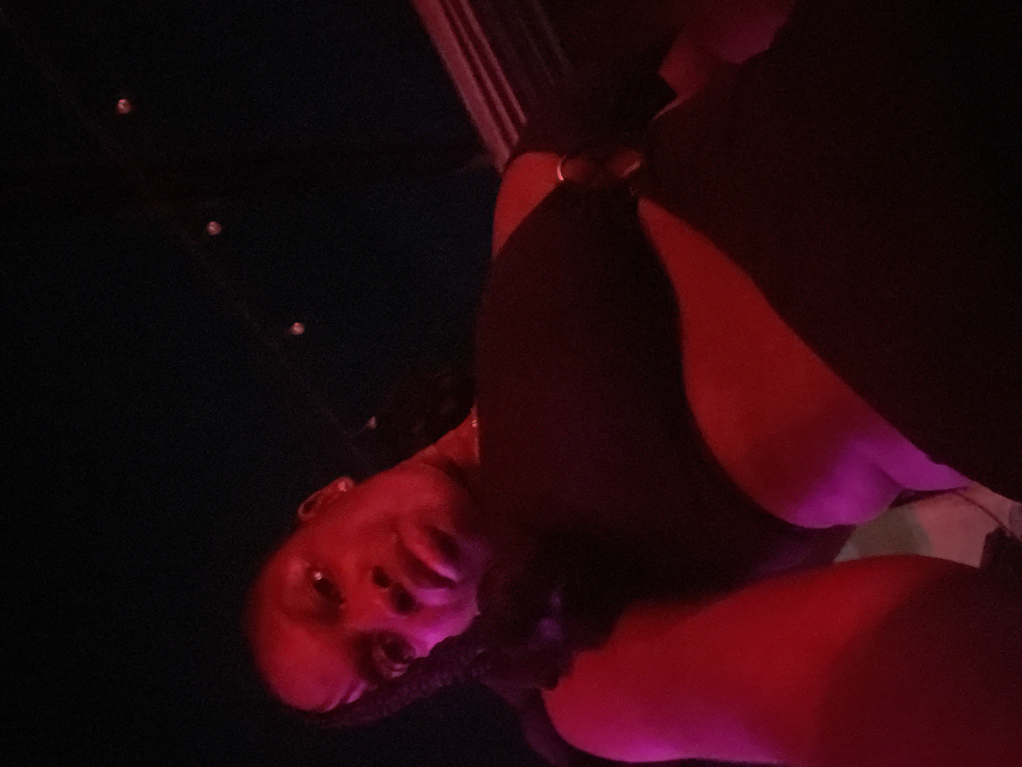 A low-angle, medium shot captures a person from the chest up, looking directly at the camera. The subject is illuminated by red and pink lighting, casting a dramatic glow on their skin. They are wearing a black top with a cut-out design and a ring detail on the side. Their hair is styled in braids. The background is dark, with a few blurry lights visible at the top. The scene appears to be indoors, possibly in a dimly lit venue. No specific details suggest the location is in Howayi, Nigeria.