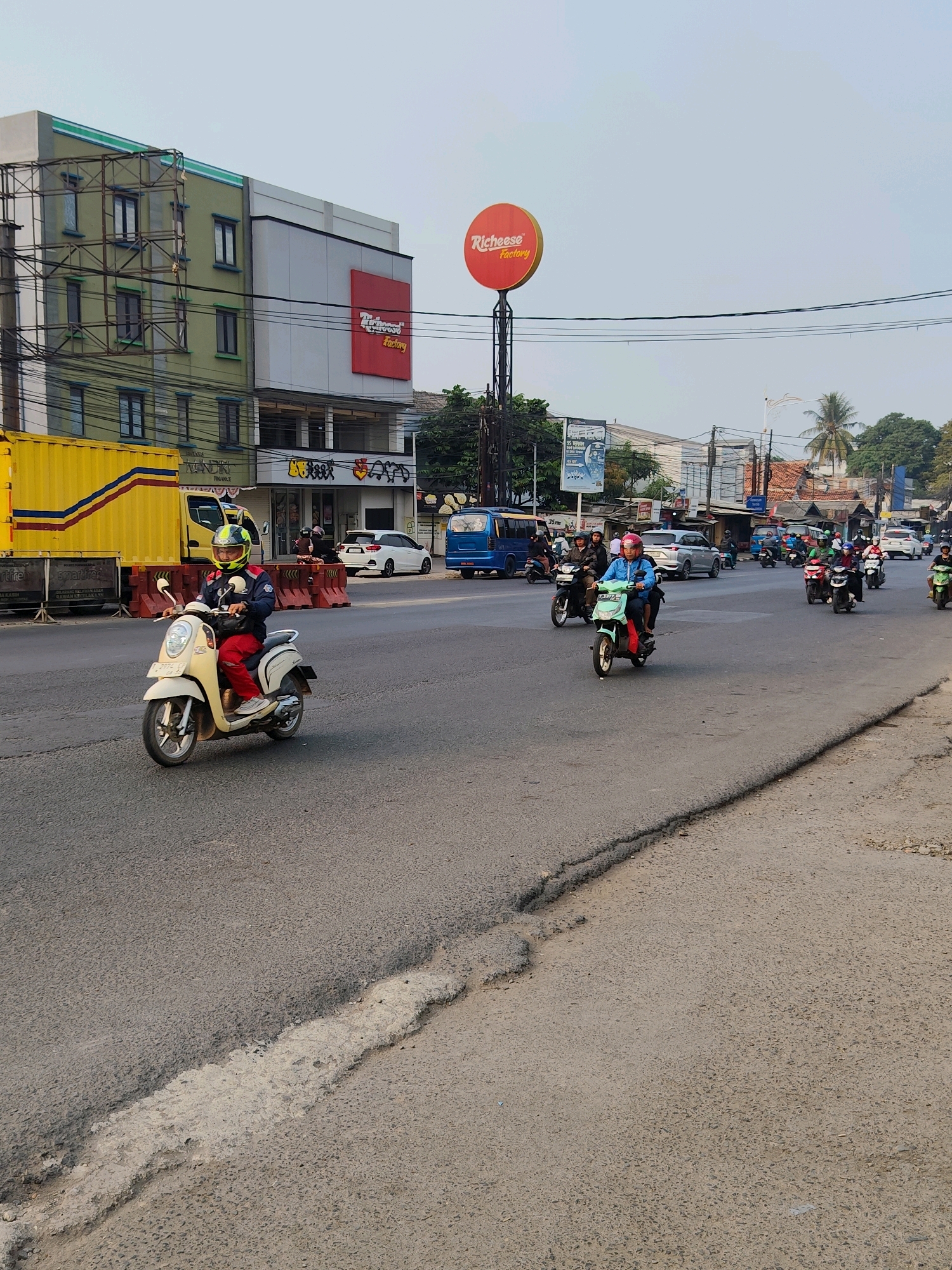 The image captures a street scene in Purwakarta, Indonesia, likely during daylight hours. In the foreground, a person wearing a blue jacket, red pants, and a yellow and green helmet rides a beige scooter. The scooter has a black bag attached to the front. Behind this scooter, a convoy of other scooters and motorcycles is visible, with riders also wearing helmets. Some riders are wearing brightly colored jackets, including green and blue. A yellow truck with red and blue stripes is parked on the left side of the road. In the background, a multi-story building with green and white facades stands, adorned with a red sign that reads "Richeese Factory." Various other buildings and trees line the street, with power lines overhead. The road itself appears to be asphalt and shows some signs of wear, with a visible crack and patch in the pavement in the foreground. Several cars and a blue bus are also present on the road, suggesting moderate traffic. The sky is clear and pale blue, indicating fair weather.