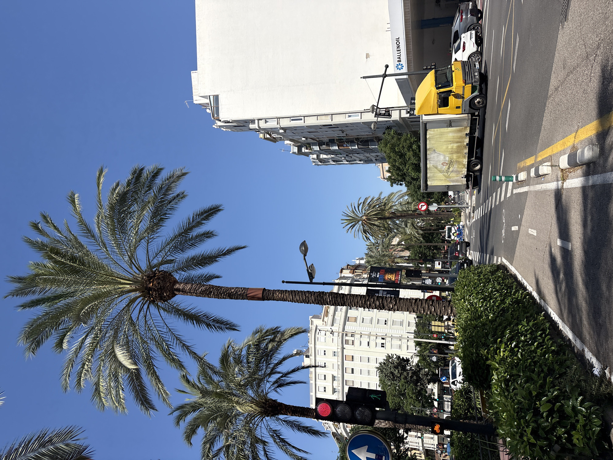 The image depicts a sunny day on a city street, likely in Valencia, Spain, judging by the palm trees and architecture. In the foreground, a lush green hedge lines a pedestrian walkway, which transitions into a road with clear lane markings. A yellow delivery truck with "Schweppes" branding on its side is driving on the right side of the road. To the right of the truck, a white car and a dark gray car are also in traffic. Further down the street, several other vehicles and pedestrians are visible, as well as buildings with balconies and awnings. A tall white building with the "BALLENOIL" logo on its facade is prominent on the right side of the image. Traffic lights are visible, with one showing a red light. Palm trees are scattered throughout the scene, some towering over the street and buildings. The sky is a clear, bright blue.