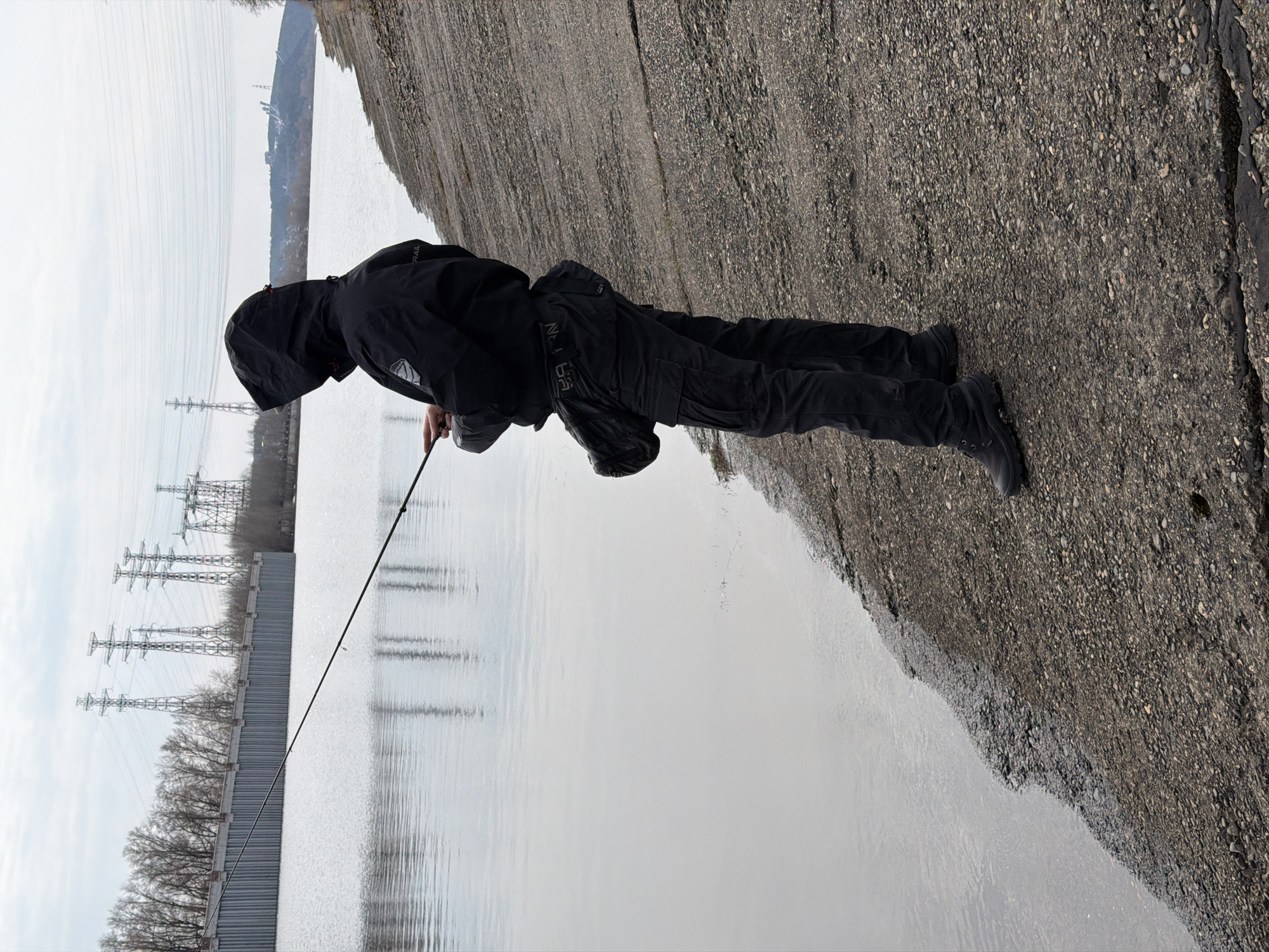 A person dressed in all black clothing, including a hooded jacket, is fishing at a body of water. The person is standing on a concrete or asphalt surface near the edge of the water, holding a fishing rod. Electrical towers are visible on the opposite side of the water behind a gray wall of vertical ribbed metal. The water appears calm and reflects the sky, which seems overcast. The concrete surface has a rough, textured appearance.
Based on the presence of electrical towers and large bodies of water, the location is likely in the city of Chaikovsky, Russia.