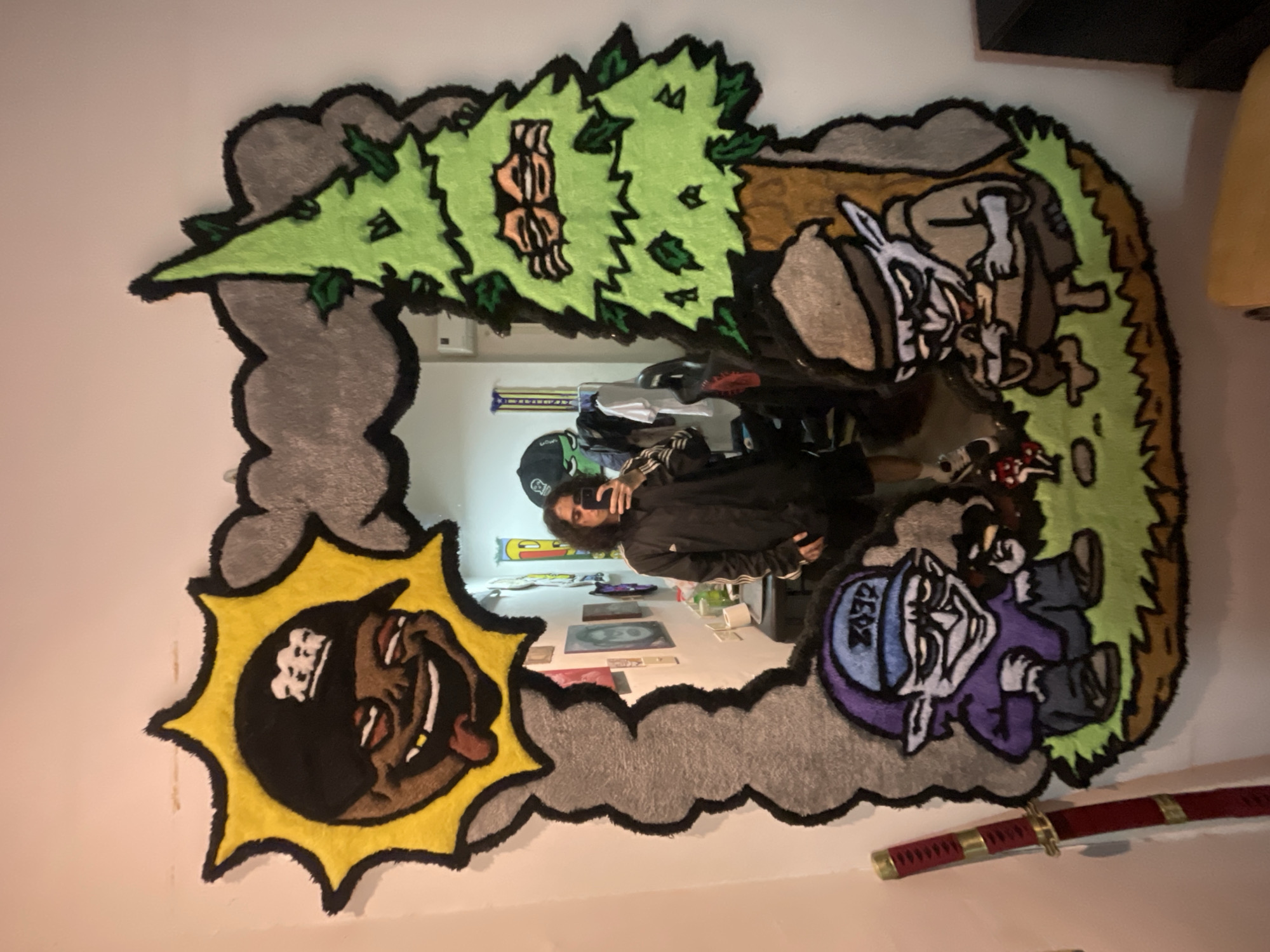 A full shot depicts a custom-designed mirror with cartoon-like rug art adhered to the frame, hung on a white wall in a room. The top left corner features a light gray cloud with a jagged edge and a green tree with what appears to be a face embedded. To the right of the tree, there is a cartoon depiction of a rabbit. At the bottom left corner, a yellow sunburst design incorporates a smiling black cartoon face wearing a black hat with a white logo. The bottom right corner displays a purple cartoon figure wearing a blue hat. Below the right corner is a red-handled katana. The mirror reflects a person taking the picture, the room’s interior, and artwork displayed on the wall behind the person. The location is not specified, and the city of Buenos Aires, Argentina, is not evident in the image.