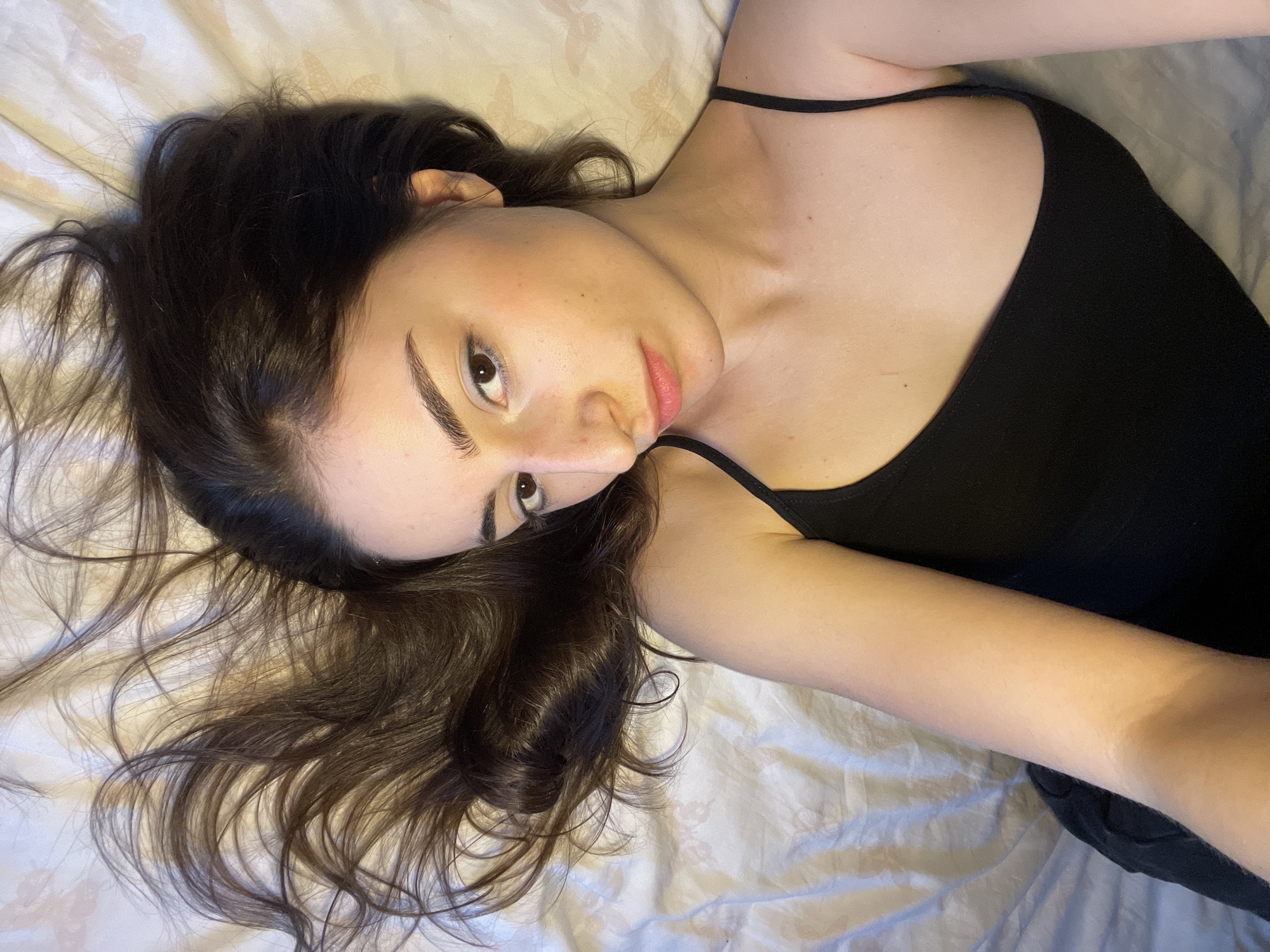 A young woman lies on a white, patterned bedsheet. She has long, dark brown hair spread around her head. She is wearing a black tank top. Her face is tilted towards the camera, and she is looking at the viewer. The background is blurred and shows the white bedsheet. The location is not discernible from the image.