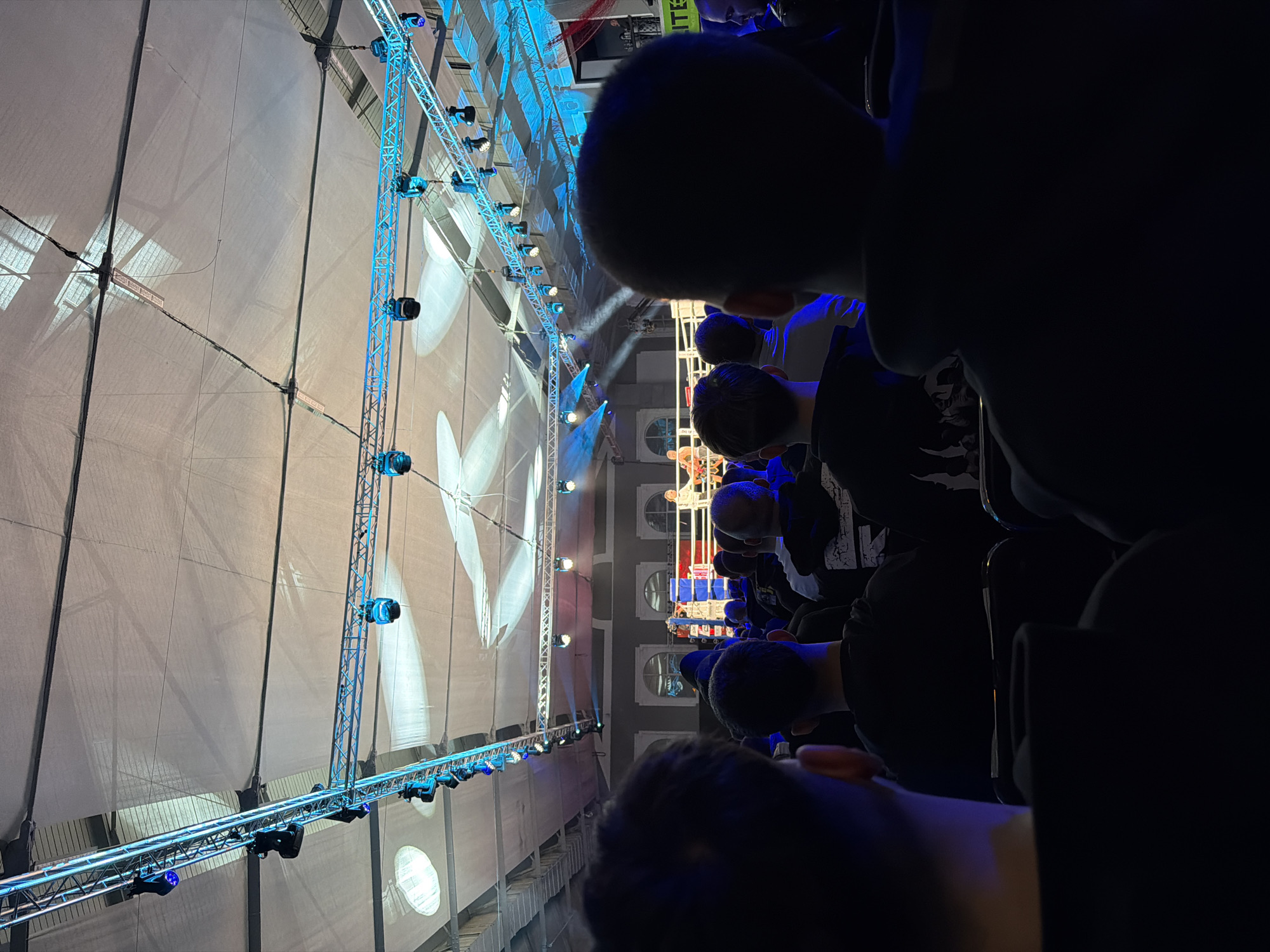 The image shows an indoor event, possibly a sporting competition, viewed from a high vantage point within a large arena. The focus is on a central stage or performance area with a complex lighting rig overhead. The lighting structure is constructed of metal beams with multiple blue-lit spotlights. A projection is displayed on a large backdrop, featuring a faint, stylized graphic.
In the foreground and side of the image, numerous people are seated, their heads and shoulders visible. Most are facing the stage, presumably watching the event. Their clothing is dark, suggesting casual attire. The background shows a wall with architectural features. The building appears to be the Arena Armeec in Sofia, Bulgaria based on the distinctive large projection screen, and architecture of the interior.