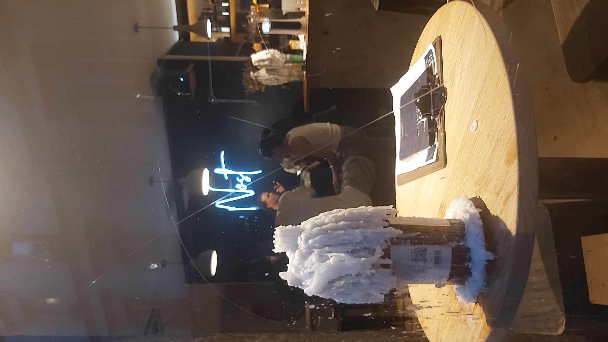 An indoor scene captured through a window. In the foreground, on a round wooden table, sits a bottle covered in melted white wax. The table also holds a clipboard with papers. In the background, through the window, two individuals are seated at a table. One person, a man, is wearing a light-colored t-shirt. The other person, a woman, is wearing a light-colored tank top. Behind them, a neon sign reads "Nost." The overall environment appears to be a bar or restaurant, likely in Paris, France. The glass in front of the scene has scratches and smudges.