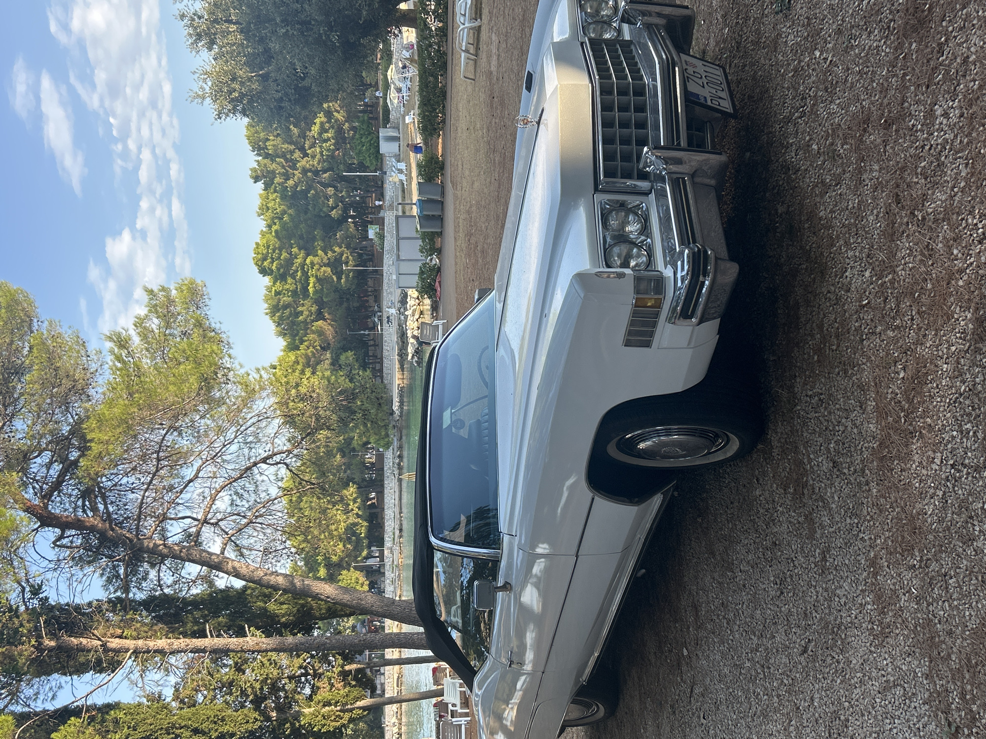 A white Cadillac convertible is parked on a gravel surface. The car is positioned at a slight angle to the left, with its front end facing forward and to the right. The car's black convertible top is up. The background shows a body of water, trees, and beach chairs, suggesting a coastal or resort location. The sky is blue with scattered white clouds. The license plate on the car reads "ZG PV-0011". This scene is located in Monterol, Croatia. There are no people actively engaged in any specific actions within the frame.