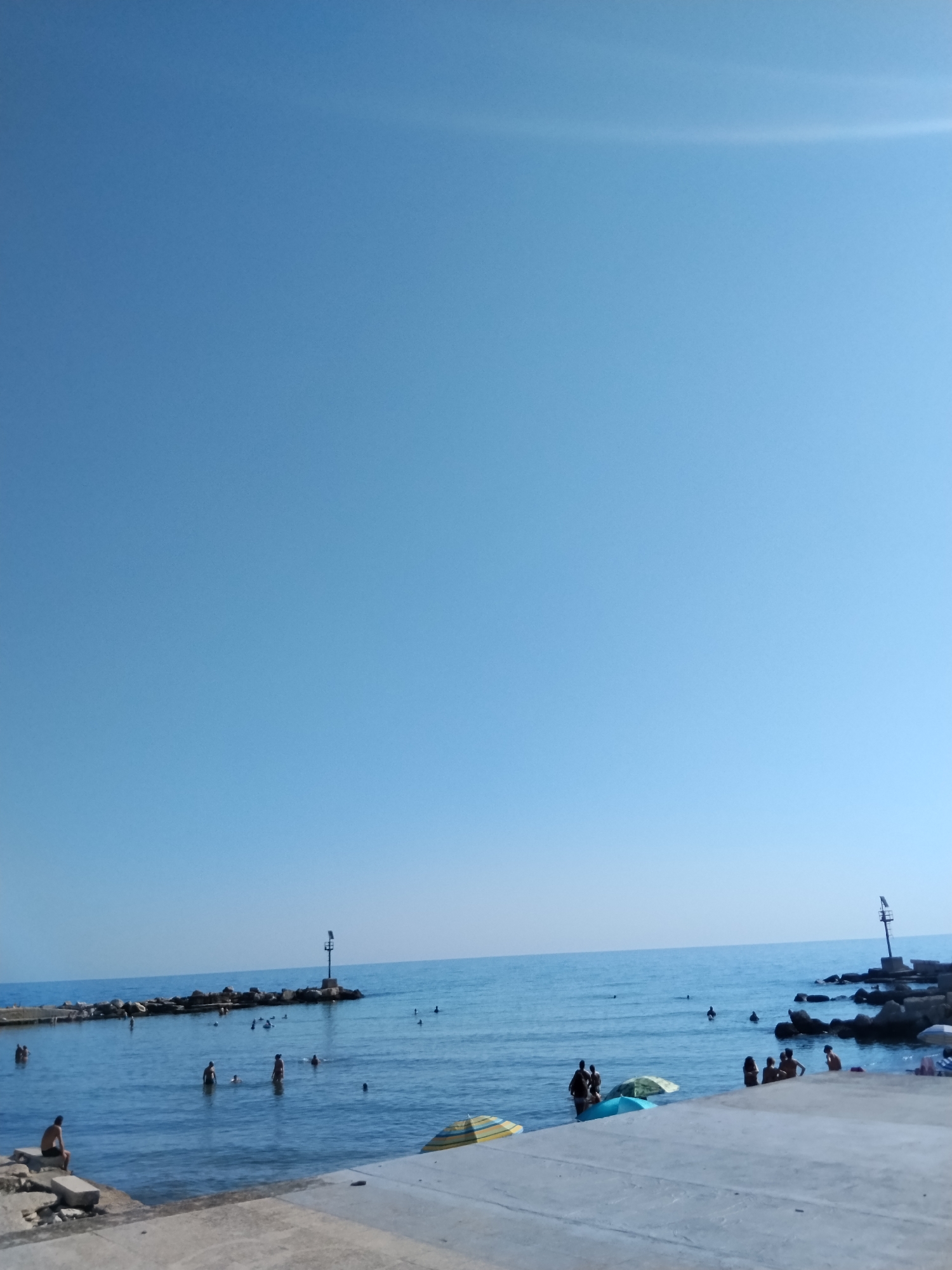 The image depicts a sunny day at a coastal area in Bari, Italy. The sky is clear and blue, with a hint of haze near the horizon. The sea is calm and a deep blue, with small waves lapping against the shore.
In the foreground, a concrete walkway lines the edge of the water. Several people are present on the walkway and in the shallow water. Some are sitting on the concrete, while others are wading or swimming. Two umbrellas, one yellow and striped and the other turquoise, are open on the walkway, providing shade.
Further out in the water, several people are swimming and enjoying the sea. A jetty made of large rocks extends into the sea, with a navigation marker on its end. Another similar jetty with a navigation marker is visible in the distance on the right side of the frame. The overall scene suggests a leisurely day at the beach, with people taking advantage of the good weather.