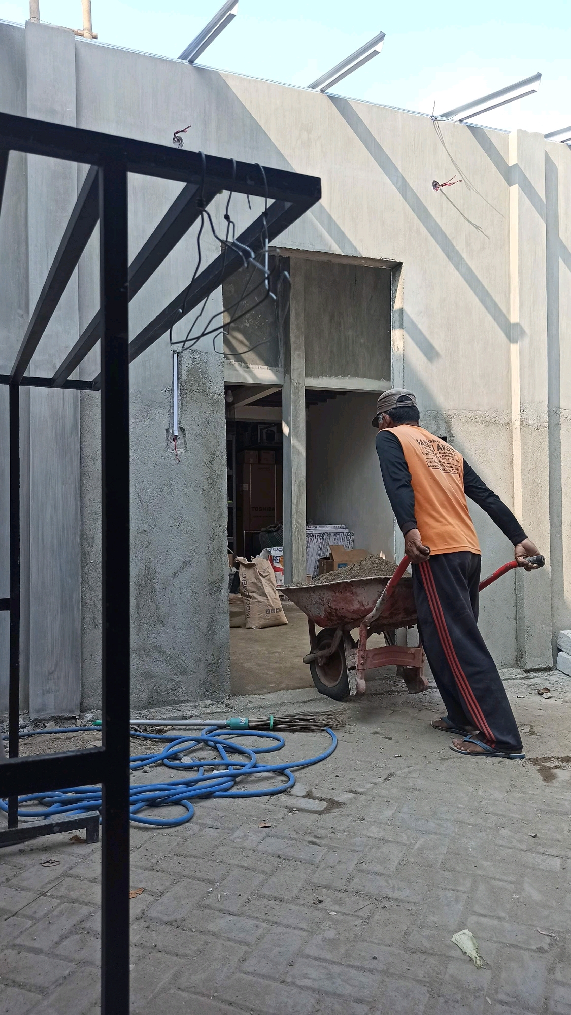 In Ngumpul, Indonesia, a man wearing an orange long-sleeved shirt and dark pants with red stripes, and a baseball cap, is pushing a wheelbarrow filled with sand. He is moving from the left side of the frame towards the right, away from an open doorway. In the foreground, a blue hose and a broom lie on the paved ground. The background shows a building under construction with concrete walls and a metal roof structure. The scene appears to be an outdoor construction site.