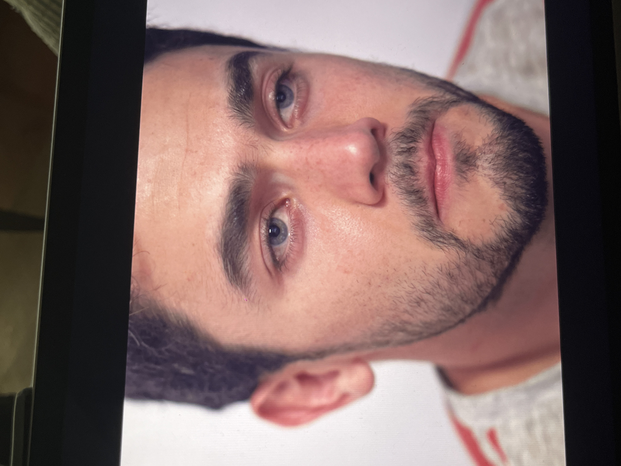 A close-up photograph shows the face of a fair-skinned man with dark hair and blue eyes. He has a short, dark beard and light stubble on his face. The skin has some visible freckles. He is wearing a white shirt with a red stripe visible near his shoulder. The photograph is oriented at an angle, with the man's face tilted towards the right. The image is displayed on a screen with a dark frame. There is no indication of a location in Mexico City.