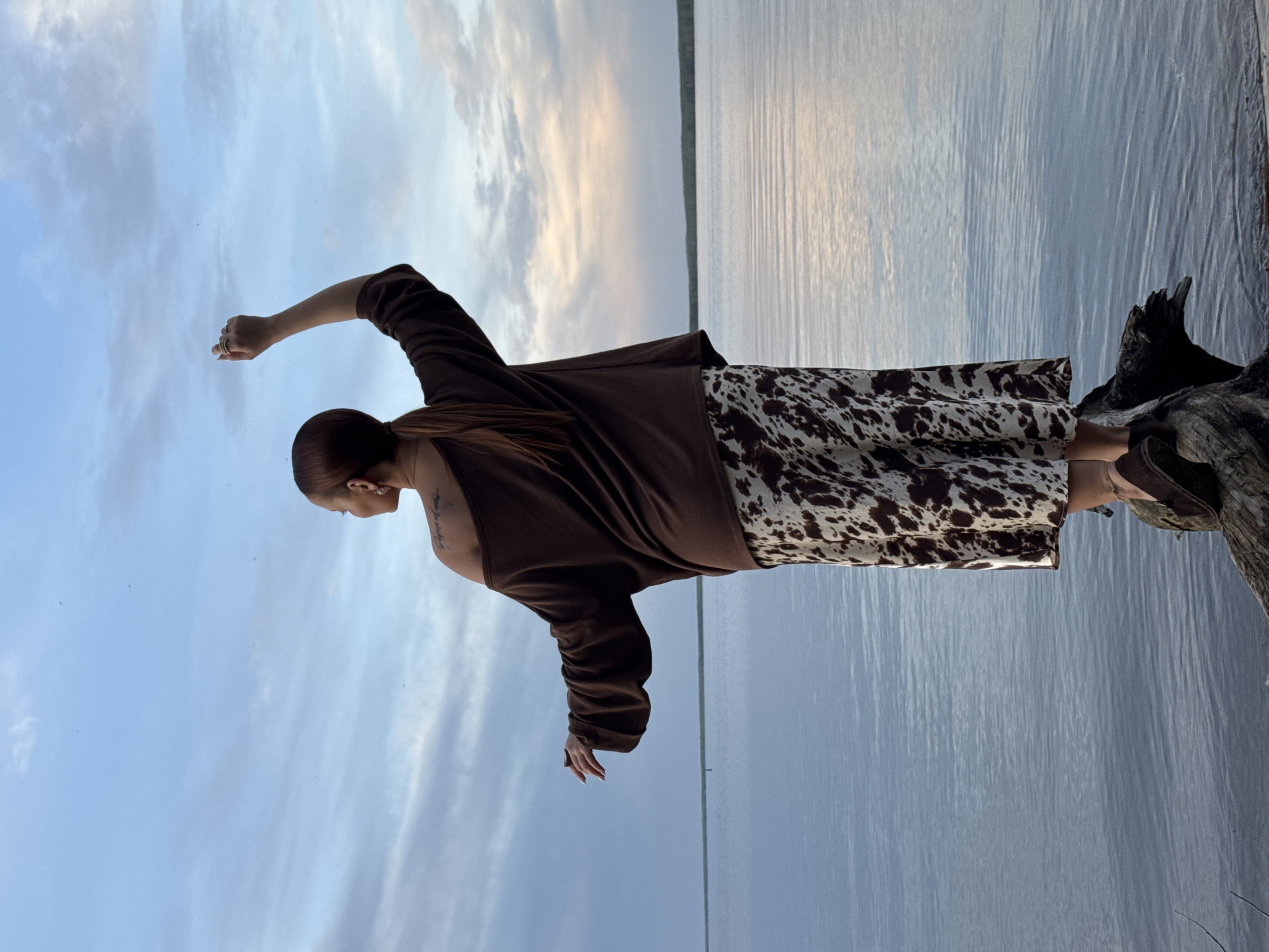A person is standing on a piece of driftwood, with a lake in the background. They are facing the water with their right arm raised towards the sky and their left arm extended towards the water. They are wearing a brown sweater and a skirt with a cow print pattern. The sky is blue with white clouds. The location is Chaikovsky, Russia.