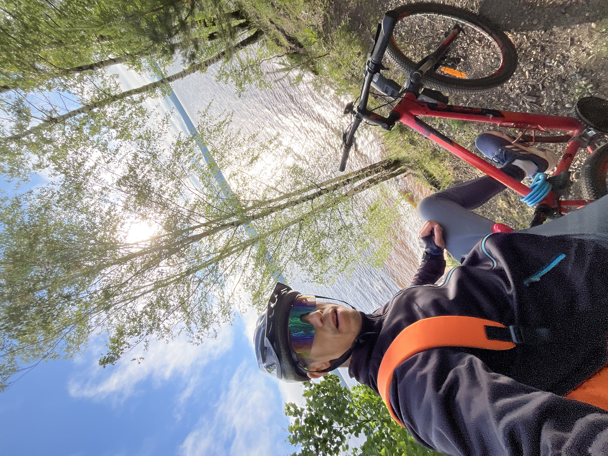 An eye-level, outdoor, medium shot captures a male cyclist in a self-portrait. He is wearing a black helmet, iridescent cycling glasses, a black zippered jacket with an orange backpack strap, grey cycling pants, and blue athletic shoes. His left arm is extended, holding the camera for the selfie. A red bicycle is parked to his right, with the handlebars and front wheel visible. The cyclist is seated on the ground on the edge of a slope overlooking a large body of water, likely the Kama River, reflecting the bright sun. The background consists of trees and a blue sky with white clouds, suggesting a sunny day. The terrain is a dirt path with small stones and scattered green foliage. The location is the embankment of the Kama River in Chaikovsky, Russia.