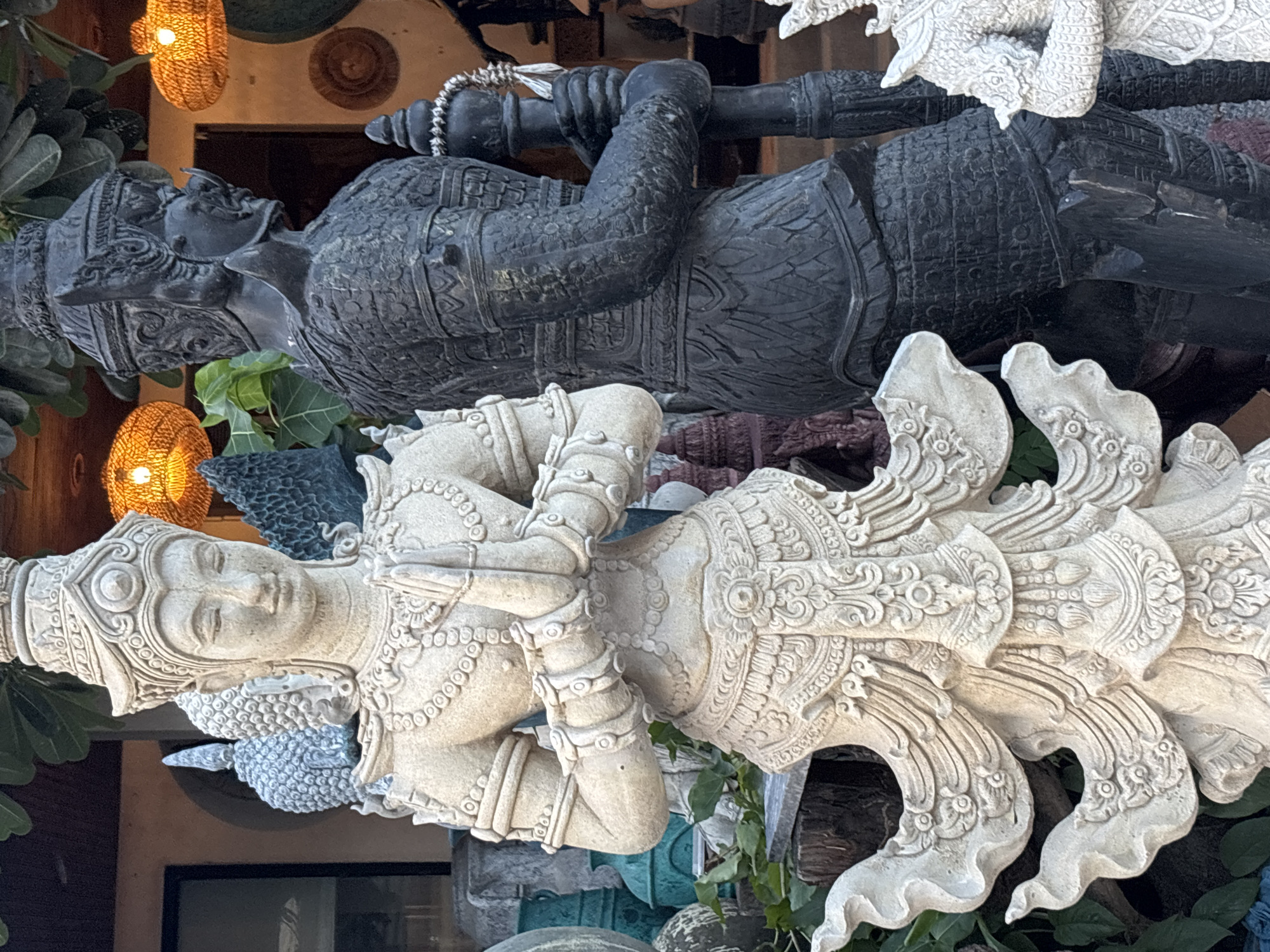 The image shows two ornate statues in Baan Bo Phut, Thailand. The top statue is dark gray, depicts a warrior with intricately detailed armor, and holds a staff. The bottom statue is light tan, representing a mythical figure with its hands pressed together in a gesture of prayer, and features elaborate wings or tail feathers. The statues are surrounded by foliage and positioned in front of a building with lit woven lanterns.