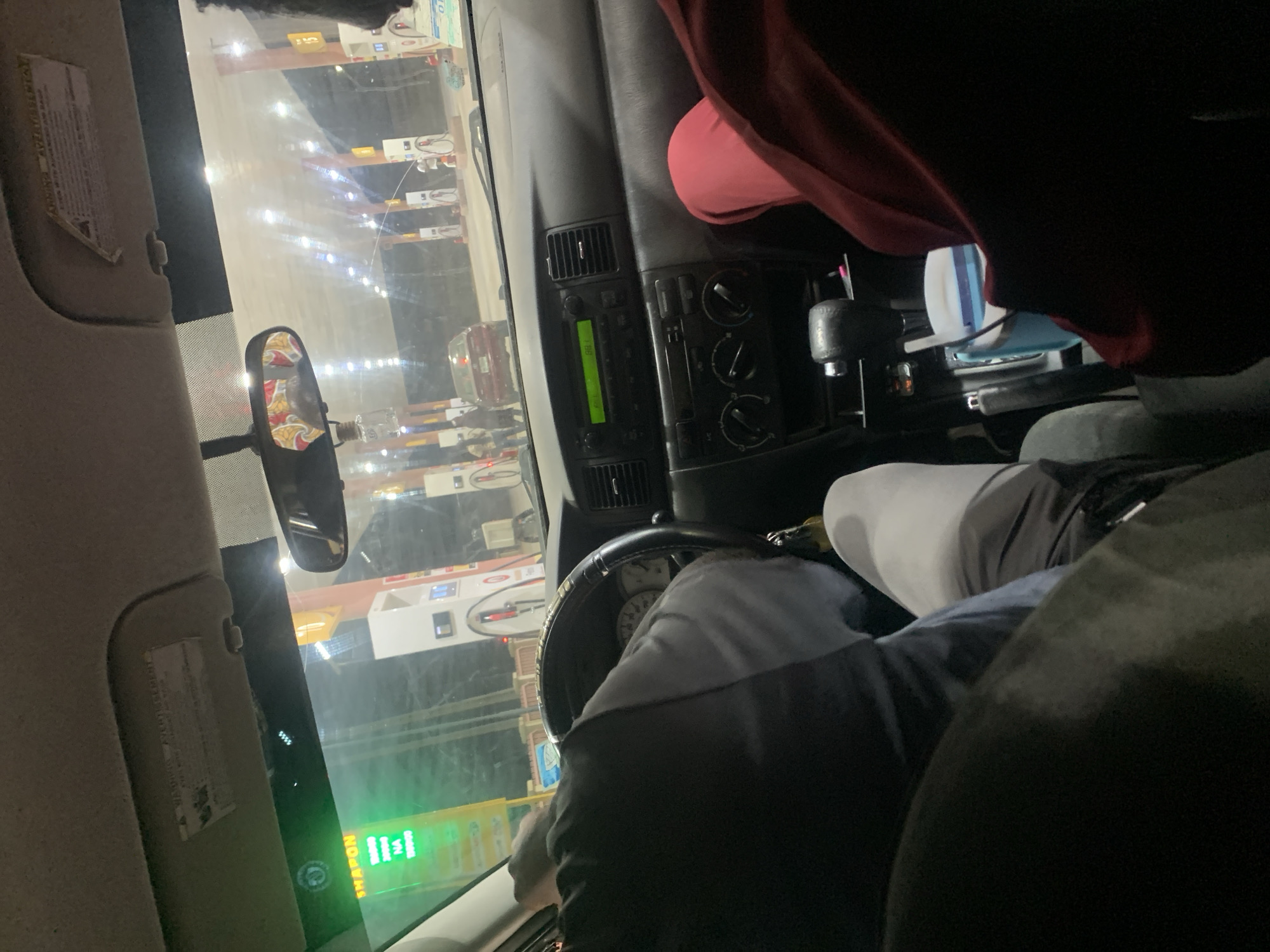 A view from the driver's seat of a car, possibly a Volkswagen, facing a gas station in Jiru, Nigeria. Multiple gas pumps are visible through the windshield. The driver is partially visible, wearing a gray and black jacket. The dashboard features a radio with a green display. The rearview mirror is visible, and the sun visor is down on the driver's side. A red garment is on the passenger side.
