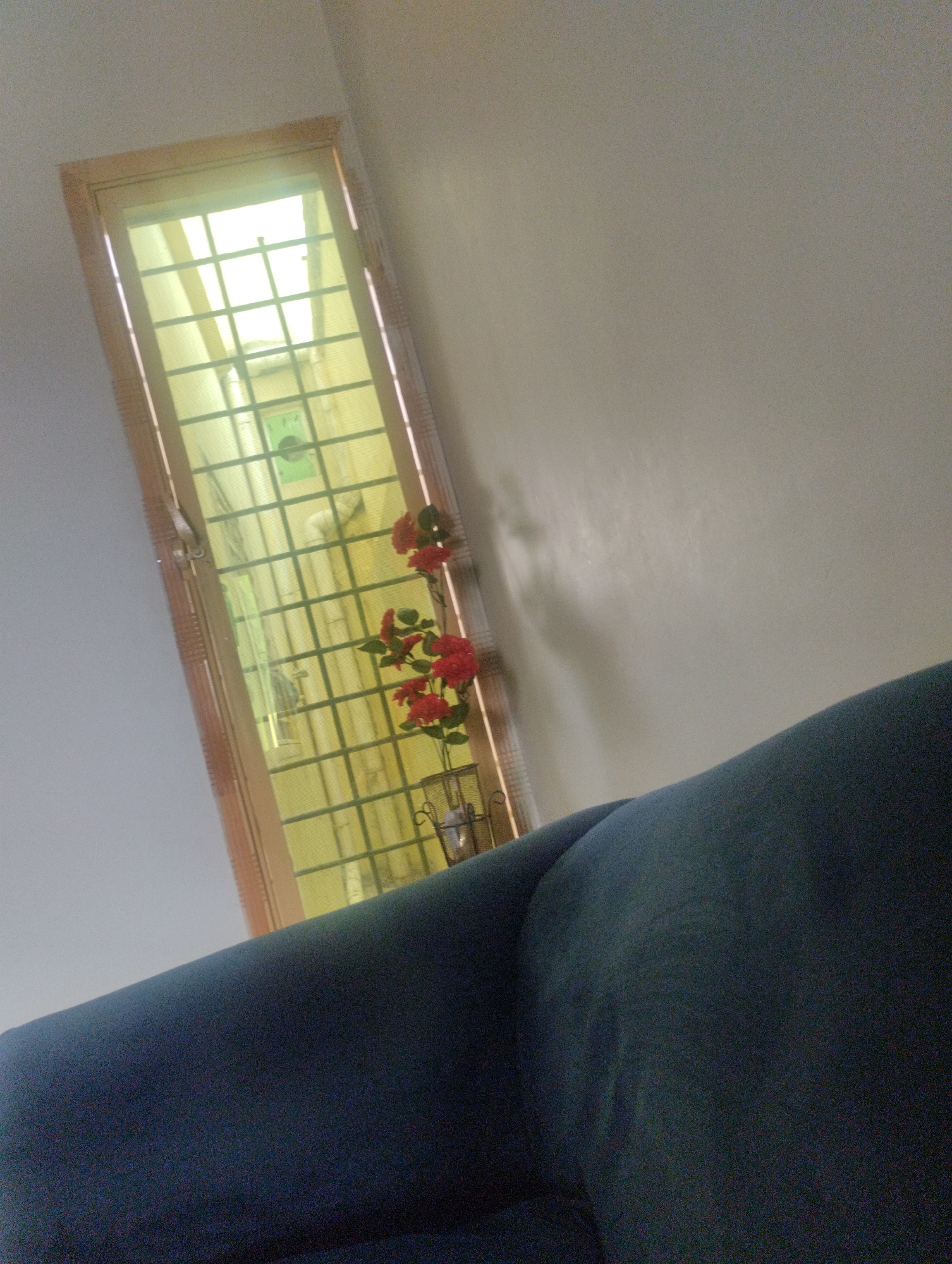 The image depicts an interior scene, likely a room, with a window and a piece of furniture. The window is tall and narrow, framed with light-colored wood. It has a grid-like pattern across it and is covered with a yellow screen or film, allowing a bright, diffused light to enter. Through the screen, indistinct shapes and a circular green object are visible, suggesting an outdoor or a different indoor space.
In front of the window, a decorative artificial plant with vibrant red roses and green leaves is placed in a dark, possibly metal, holder. The roses are the most prominent splashes of color in the image.
In the foreground, a large portion of a dark blue upholstered furniture item, likely a sofa or chair, occupies the lower half of the frame. The furniture's texture is visible, with slight creasing suggesting it is made of fabric. The camera angle is tilted, giving a dynamic, slightly off-kilter perspective of the scene.
The lighting suggests it is daytime, with the bright light coming through the window. The overall atmosphere is still and quiet, with no people or discernible activity. The setting appears to be domestic, possibly a living room or a sunroom. There is no visible text in the image.