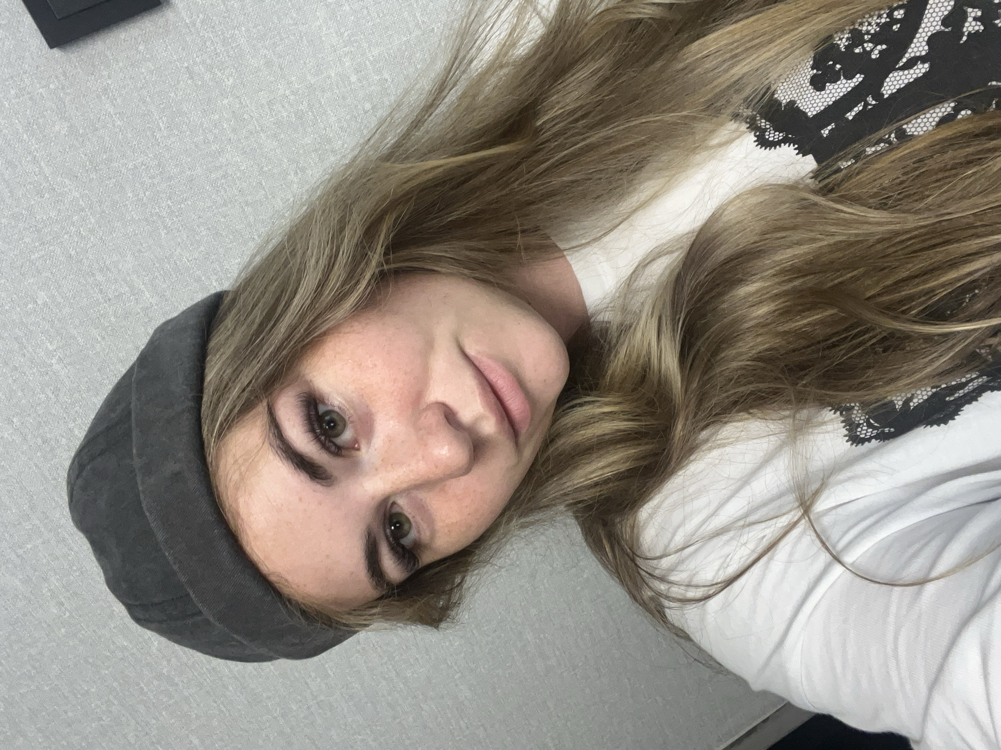 A young woman with blonde hair and freckles wears a grey beanie and a white t-shirt with a black lace design. She is indoors, possibly in an apartment or hotel room, as indicated by the textured wallpaper. The lighting suggests an indoor setting, possibly illuminated by artificial light. No specific location in Chaikovsky, Russia, can be identified from the image.