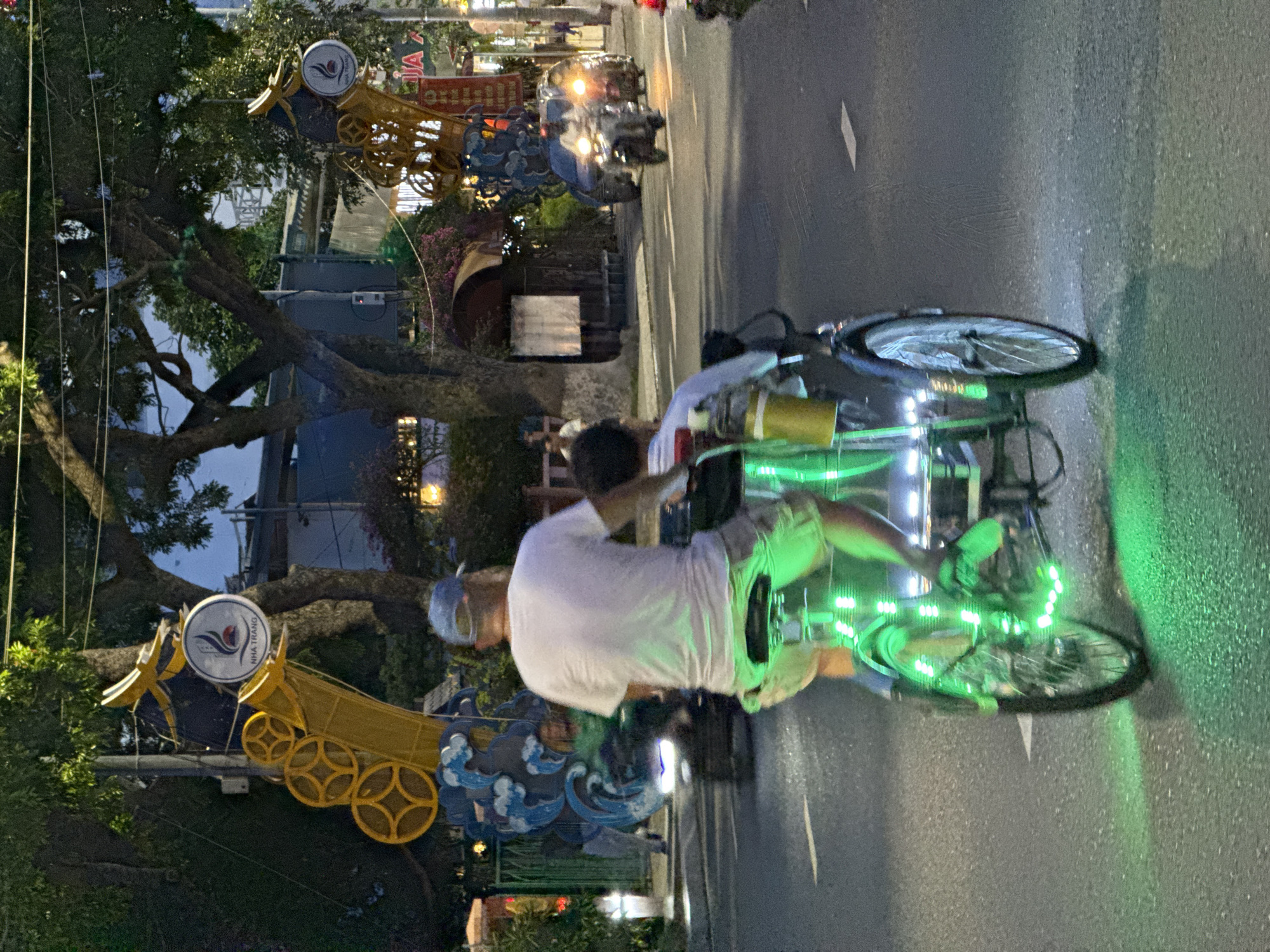 This image captures a nighttime street scene in Nha Trang, Vietnam. In the foreground, a man wearing a white t-shirt and a blue cap is riding a cyclos, a three-wheeled vehicle, which is illuminated with bright green LED lights. A child is seated behind him. The cyclos is moving along a paved street, with white lane markings visible. In the background, there are trees with decorative yellow structures and circular motifs, possibly part of a street decoration or a building facade, bearing a circular logo with "NHA TRANG" written on it. Further back, other vehicles, including motorcycles, are visible, some with illuminated headlights. The overall atmosphere is one of a busy urban environment at night, illuminated by artificial lights.
