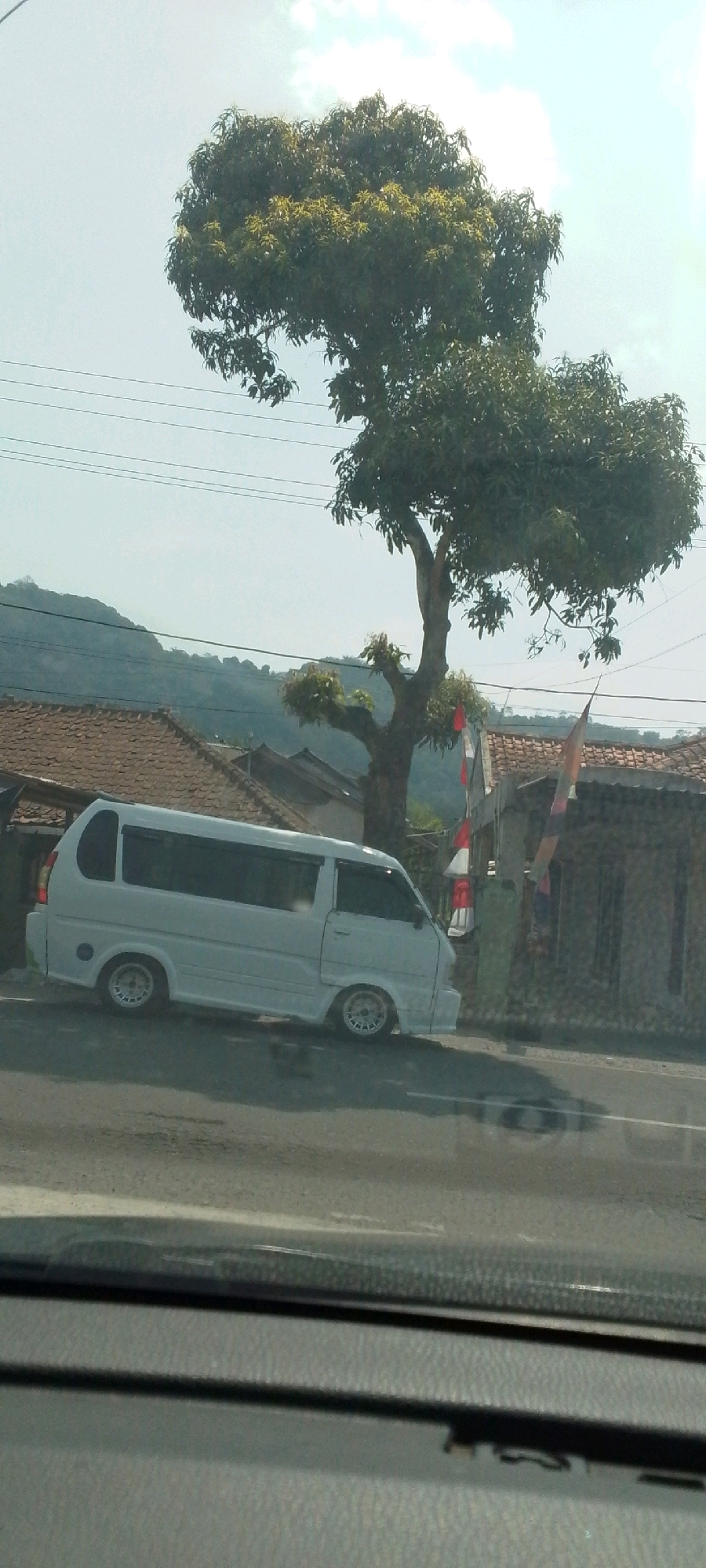 A white van is parked on the side of a road, with a tree and houses in the background. The van is facing left and appears to be in good condition. The houses have red tiled roofs and one has a flag with red and white stripes. The tree is large and leafy, with some branches trimmed. The sky is bright and overcast, with some clouds visible. The road is asphalt and appears to be wet, with reflections of the surroundings visible. The image was taken in Susukan, Indonesia, as indicated by the presence of Indonesian flags.