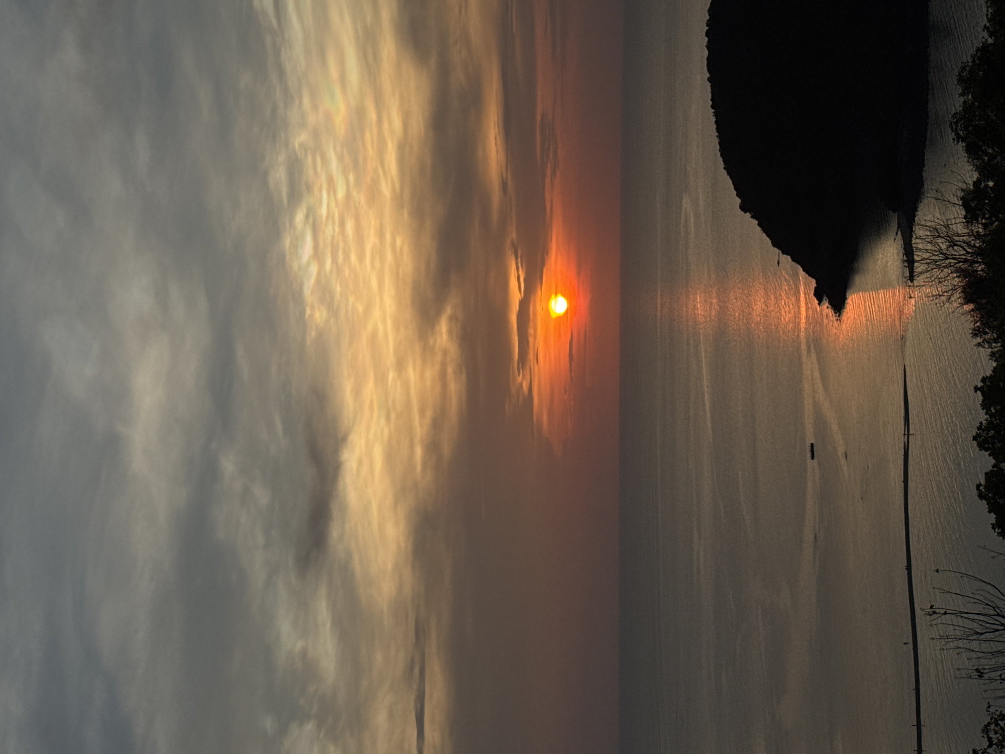 The image shows a sunset over water in Baan Wang Ta Kien, Thailand. The sun is low on the horizon, creating a bright orange glow. The sky has both dark and light clouds, with the sun breaking through some of the lighter clouds. Part of an island with trees on it is visible on the right side of the image. In the water, there are what appear to be fishing nets. A small boat is also visible in the water.