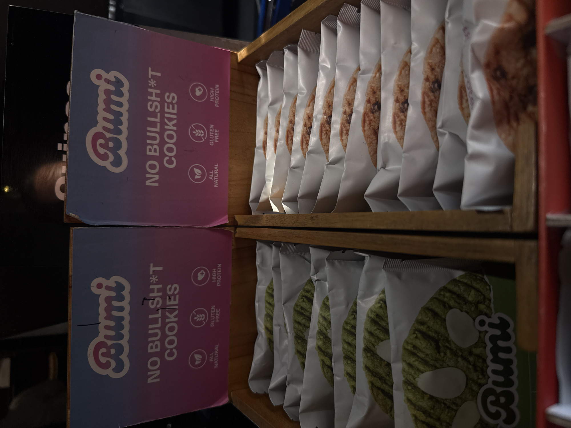 This media file captures a retail display featuring "Bumi NO BULLSH*T COOKIES" in an indoor setting, likely a store or market. The image is taken at an angle, showing two rows of packaged cookies neatly arranged in wooden display shelves.
On the left, two prominent promotional cards or boxes display the brand name "Bumi" in a cursive font. Above the top shelf, one card features a pink-to-blue gradient background, while a similar card below the bottom shelf has a green background. Both cards prominently display the text "NO BULLSH*T COOKIES" and list product attributes with icons: "HIGH PROTEIN," "GLUTEN FREE," and "ALL NATURAL."
The top shelf contains cookies that appear to be chocolate chip, individually packaged in white or light-colored wrappers, showing parts of the cookie through transparent sections. The bottom shelf holds a different variety of cookies, green in color with visible white almond slivers, also individually packaged in similar white wrappers, with the "Bumi" logo visible on some. The overall arrangement suggests the products are ready for purchase.