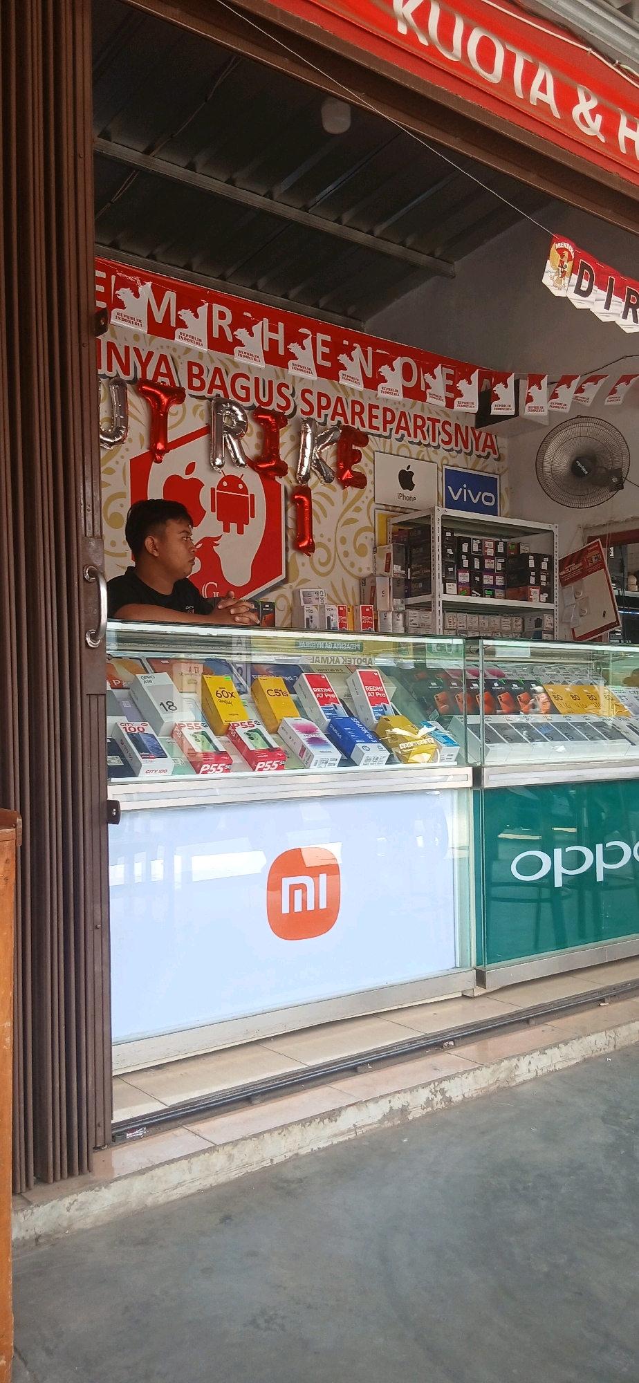 This is an eye-level medium shot capturing the storefront of a mobile phone and accessory shop in Metro, Indonesia. The shop is open, with a brown accordion-style gate partially pulled back on the left, revealing the interior. A young man with short black hair, wearing a black t-shirt, is seated behind a counter, looking to his left.
The shop's interior is brightly lit. Above the counter, the wall features a large red and white banner with Indonesian text and small red and white Indonesian flags ("Merdeka"). In front of the banner, silver and red balloons spell out "ITRIKE". A large red illustration of the Android robot logo is visible to the left of the balloons, and an Apple iPhone logo is to the right. Behind the man, shelves are stocked with numerous boxes of mobile phones and accessories. Brands like Vivo are visible on signs and product packaging.
A glass display counter in front of the man showcases rows of mobile phone boxes, with brands such as Oppo, Xiaomi (Mi logo prominently displayed on a white panel), and Redmi visible. Further to the right, a green panel displays the "oppo" logo in white text. The shop appears to be on a street, with a gray concrete ground and curb visible in the foreground. The overall impression is a busy retail environment focused on mobile technology.