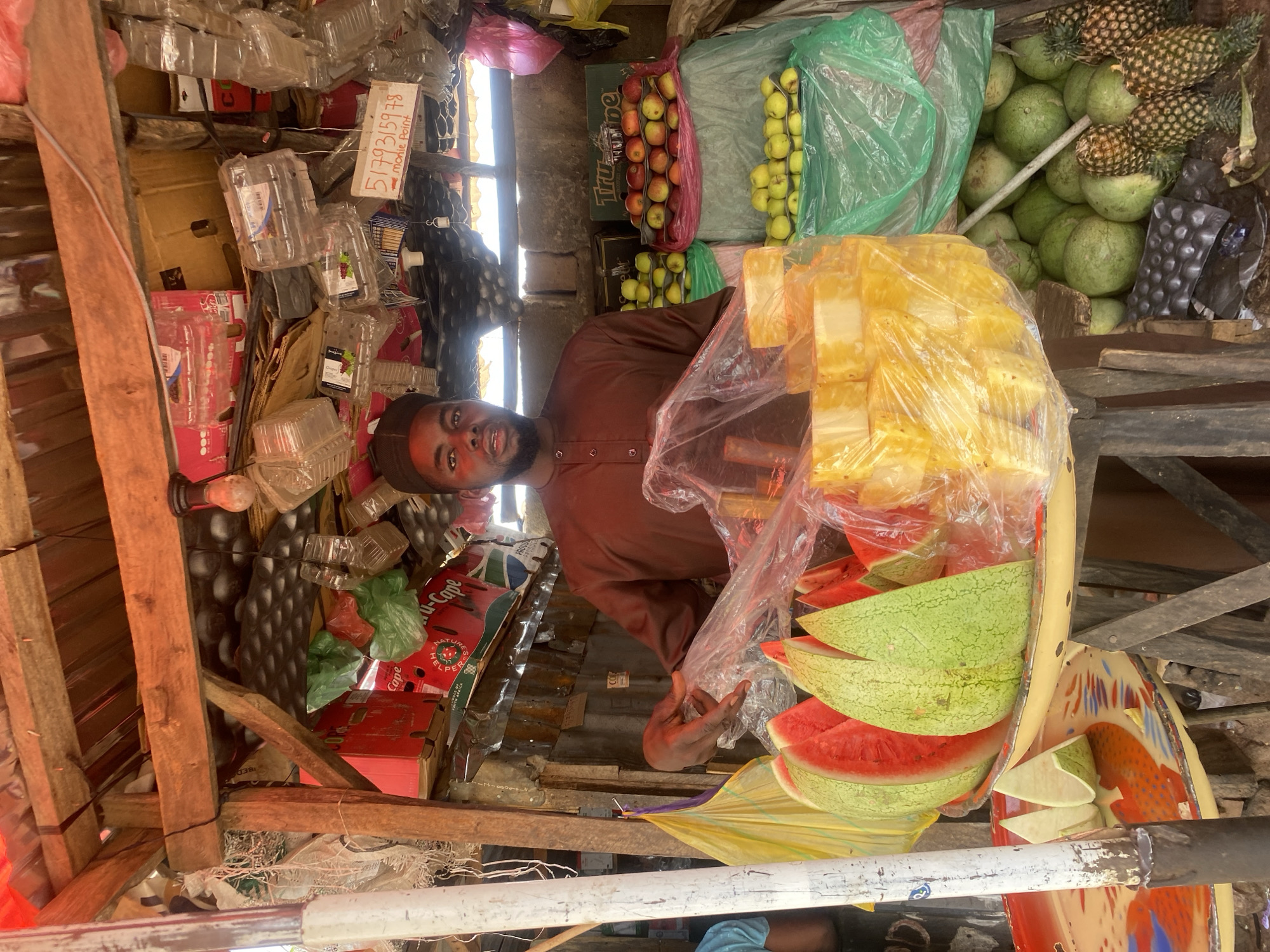 A male vendor operates a fruit stall in Sabon Tasha, Nigeria. The individual, appearing to be an adult male with dark skin and a beard, wears a dark brown long-sleeved tunic and a dark cap. He stands centrally behind a fruit display, gazing directly at the viewer. His right hand is seen holding a clear plastic bag over a quantity of cubed pineapple pieces. To his left, multiple wedge-cut watermelon slices, displaying red flesh and green rind, are arranged on a circular tray with red, orange, and blue patterns. Behind the vendor, stacked arrays of green apples and red/yellow apples are visible, with product boxes such as "Tru-type" and "Cape" in the background. The stall features a rustic construction with exposed wooden beams, planks, and corrugated metal sheeting forming the roof and rear wall. Various empty plastic containers, colored plastic bags, and cardboard boxes are suspended from the ceiling and frame. A handwritten sign, displaying "5179315978 morile Point," hangs above. On the lower right, a collection of whole green, round fruits and several whole pineapples are arranged on the ground. A white-painted pole stands vertically in the left foreground, partially obscuring an individual's arm in a blue garment. The environment is brightly illuminated.