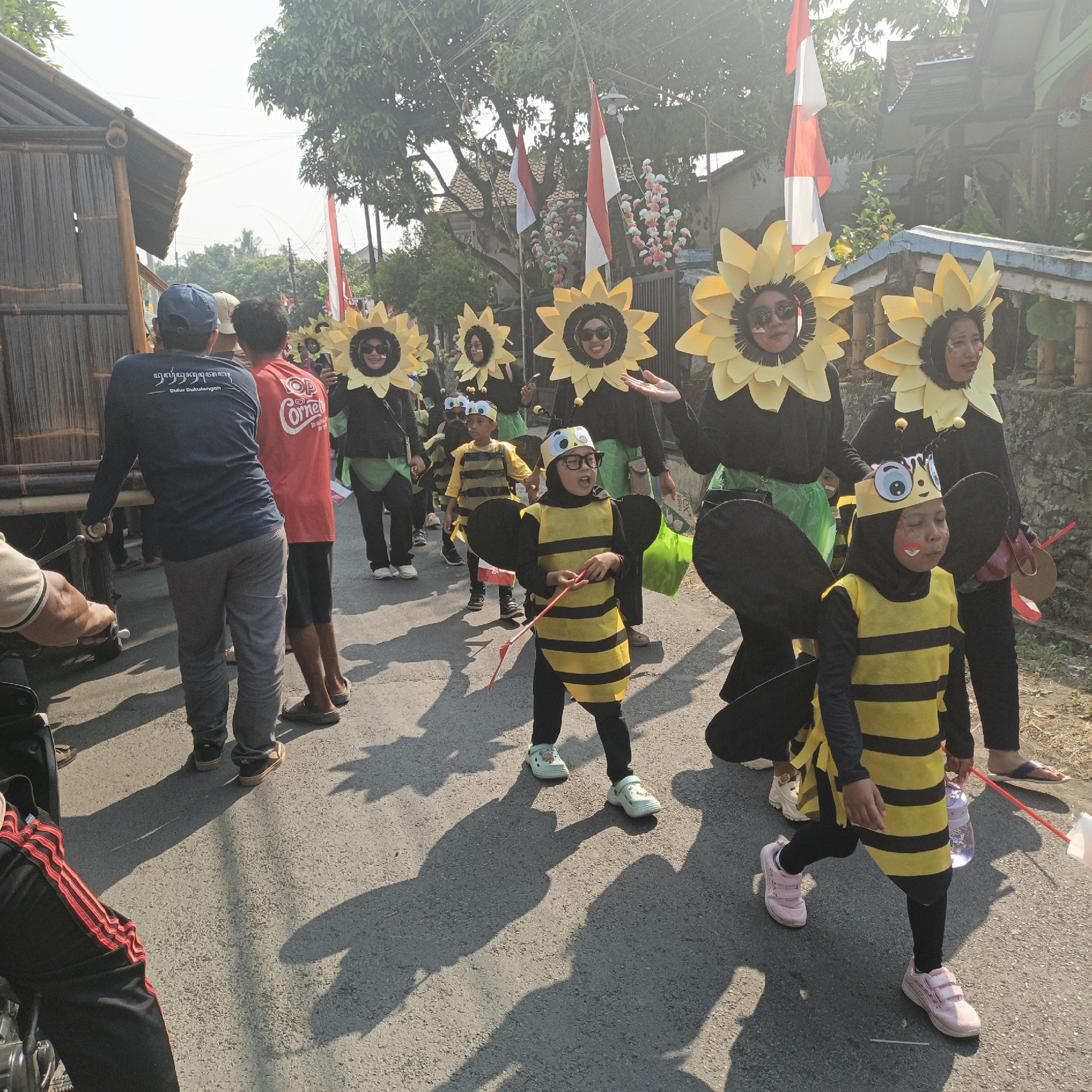 In the city of Tanahbaya, Indonesia, a festive parade is underway on a paved street under a clear, sunny sky. The procession features several individuals in elaborate costumes. At the forefront, a group of children are dressed as bees, complete with yellow and black striped outfits, antennae headbands, and attached wings. One child in the foreground, wearing glasses, holds a red stick with two streamers, while another, further back, sports face paint and a similar costume. Behind the bee-clad children, adults are costumed as sunflowers, with large yellow petals forming a halo around their heads. They wear black tops and some have green accents. One adult, in the center of the sunflower group, appears to be leading or interacting with the children. Indonesian flags are visible in the background, adding to the celebratory atmosphere. On the left side of the street, a bamboo structure suggests local architecture. The street is lined with spectators and participants, casting long shadows on the asphalt, indicating the time of day is likely late morning or early afternoon. The overall scene depicts a vibrant cultural event characterized by costumes, community participation, and national pride.