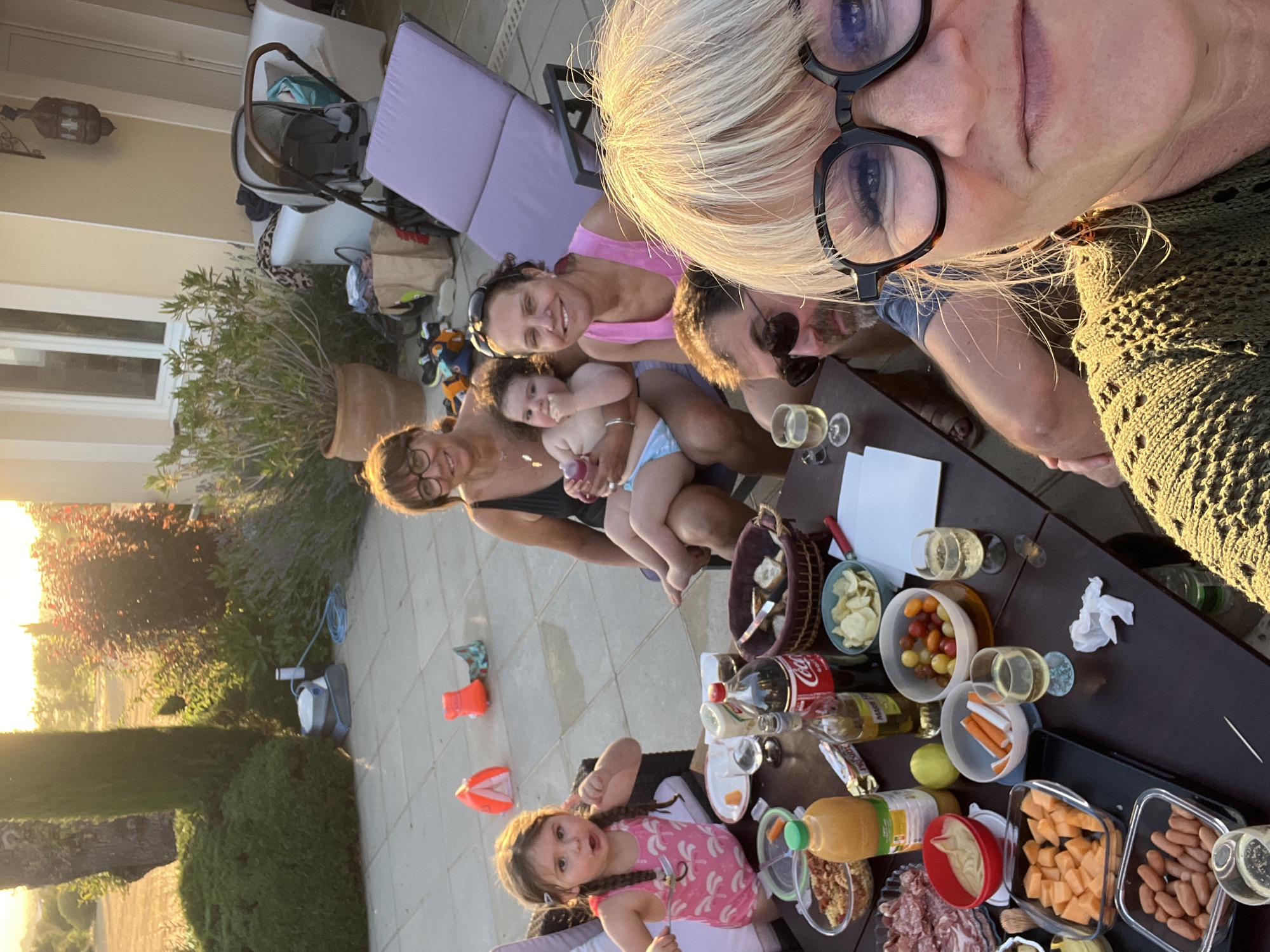 A group of people are gathered around a table outside on a patio at sunset. The patio is paved with stone tiles and surrounded by landscaping. In the background, there is a house and a stroller. On the table, there is food and drinks, including a bottle of Coca-Cola, a bottle of olive oil, chips, and various bowls of food. The people include a woman in the foreground taking a selfie, a man with sunglasses behind her, two women and a baby sitting on chairs, and a young girl eating at the table. One of the women is wearing a black swimsuit, and the baby is wearing a blue diaper. The young girl is wearing a pink swimsuit with palm tree prints. The sun is setting, casting a warm glow over the scene. The location is Saint-Galmier, France, as indicated by the context of the request.
