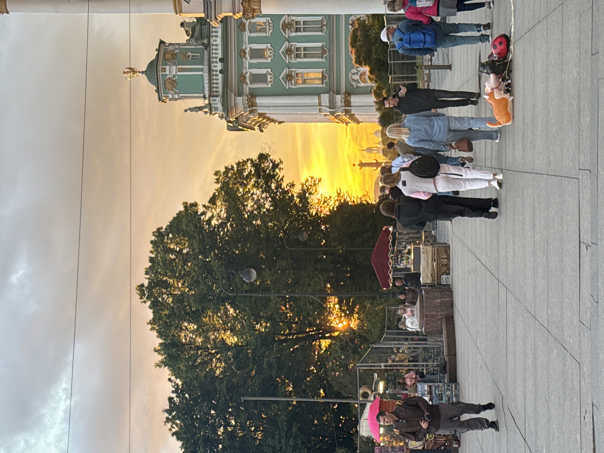 The image captures a bustling outdoor scene in Saint Petersburg, Russia, likely during the late afternoon or early evening given the warm glow of the setting sun. The sky is a dramatic canvas of orange and yellow hues, indicating a sunset that illuminates the grand architecture in the background. A large, ornate building, possibly a museum or palace, with green and white facades and numerous windows, dominates the upper left portion of the frame.
In the foreground and mid-ground, a crowd of people are strolling along a wide, paved plaza. They are engaged in various activities, from walking and talking to browsing what appear to be market stalls set up along the path. Among the people are families and individuals, some holding hands, suggesting a leisurely outing. A few individuals are holding balloons, including a distinctive ladybug-shaped one, adding a touch of festivity to the scene.
Trees with lush green foliage are interspersed throughout the plaza, providing shade and contrast to the urban landscape. The lighting from the setting sun filters through the trees, creating dappled patterns on the ground and highlighting the warm colors of the sky.
Notable details include the presence of several market stands, some with awnings, selling various goods. A bright pink umbrella is visible in the lower part of the image, held by a man who appears to be taking a photograph. The overall atmosphere seems relaxed and vibrant, with people enjoying the pleasant weather and the picturesque surroundings. The stone paving of the plaza and the classic architectural style of the buildings strongly suggest an iconic location in Saint Petersburg. No visible text is discernible in the image.