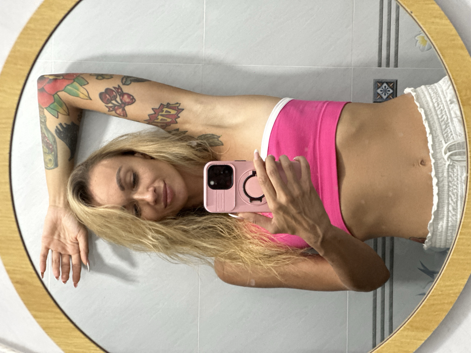 A woman with blonde hair is taking a mirror selfie in a bathroom in Mũi Né, Vietnam. She is wearing a pink bandeau top and white drawstring shorts. She has tattoos on her arm and her stomach is visible. She is holding a pink phone in her right hand, angled towards the mirror. Her left arm is raised and resting on the mirror frame. The bathroom has tiled walls and a light fixture visible in the reflection.