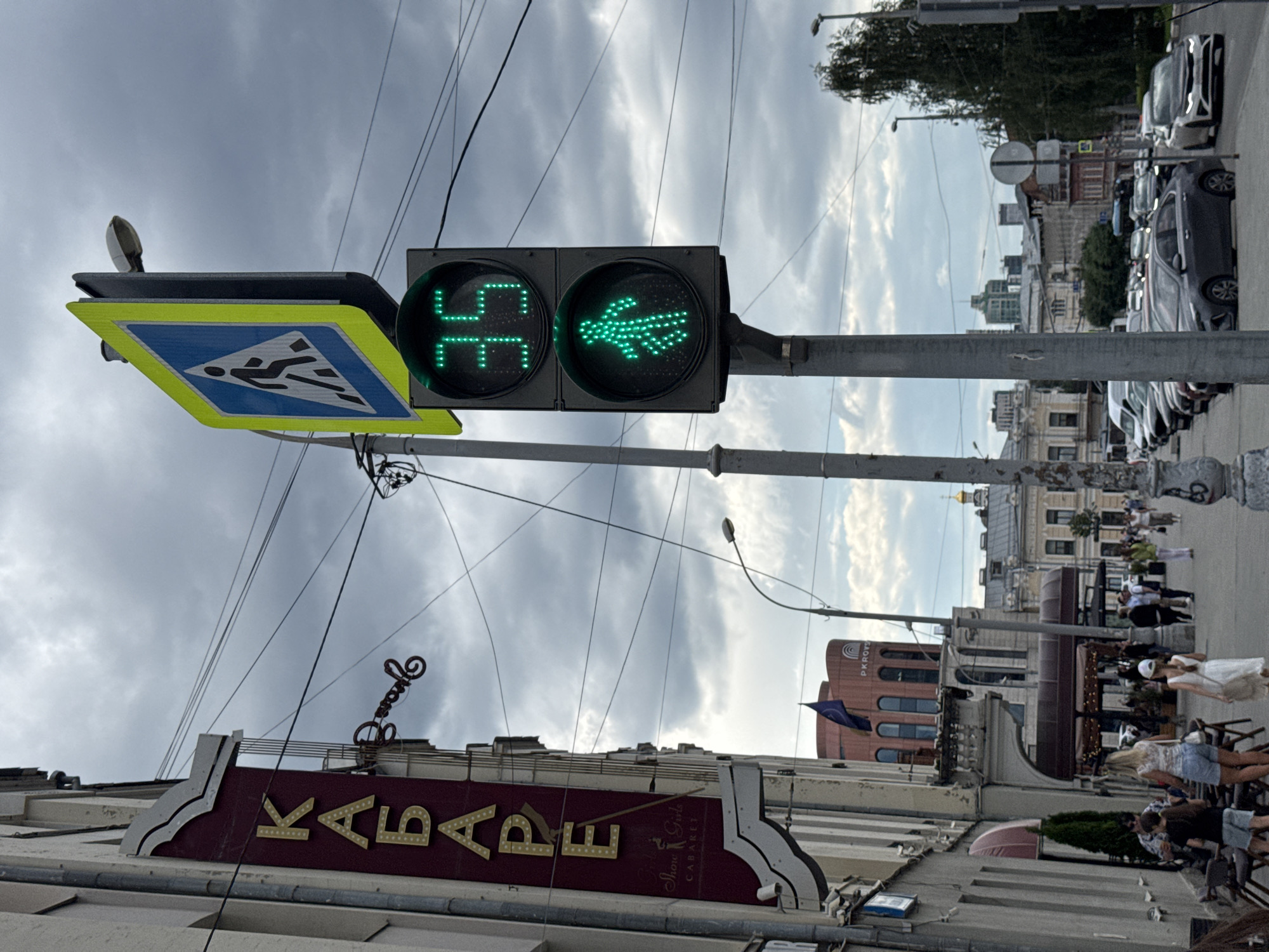 This is a street-level, medium shot of a cloudy day in Yekaterinburg, Russia, specifically on Vaynera Street, a pedestrian zone. The foreground is dominated by a traffic light pole with a pedestrian crossing sign and a traffic signal. The traffic signal displays a green walking figure and a countdown timer showing "35" seconds, indicating it is safe for pedestrians to cross. To the left of the traffic light, a tall, dark red sign with gold lettering reading "КАБАРЕ" (Cabaret) is affixed to a building. Below "КАБАРЕ," smaller text reads "Show Girls" and "CABARET."
The mid-ground shows a sidewalk with several people walking and sitting at outdoor tables. Among them are a woman in a white top and denim shorts, a young girl in a white dress, and other individuals further down the street. The street itself is lined with parked cars. In the background, buildings of varying architectural styles are visible, including a distinctive building with a curved facade displaying "PKROVS." The sky is overcast with heavy clouds. The overall impression is a busy urban street scene on a potentially cooler day.