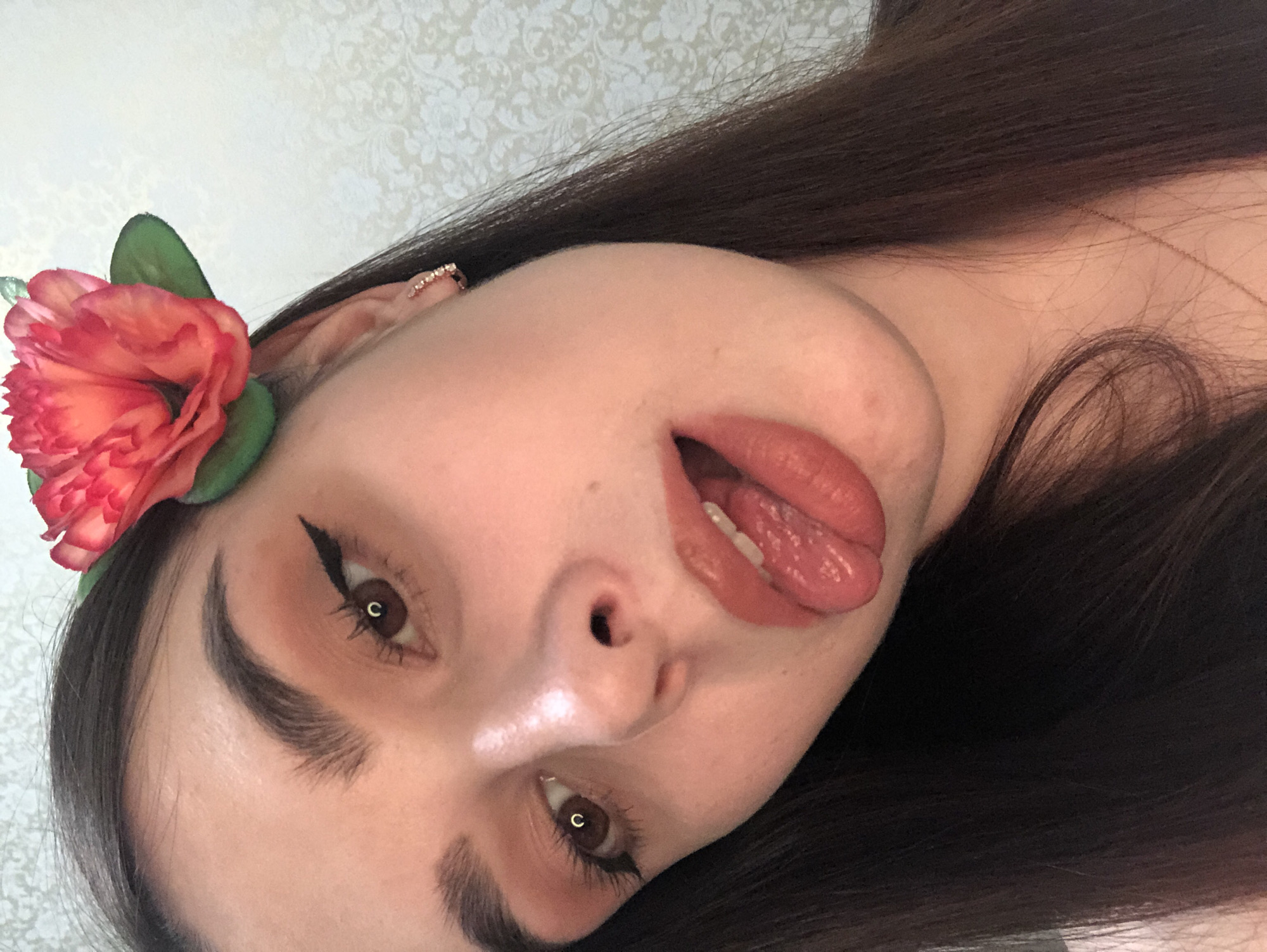A close-up, tilted selfie of a young woman. She has long, dark brown hair, fair skin, and brown eyes with winged eyeliner. She is wearing a large, artificial orange flower with green leaves near her ear. Her lips are glossed and slightly parted, revealing the tip of her tongue. She appears to have an earring in her ear. The background is a blurred, patterned wall. The location of this image cannot be deduced from the picture.