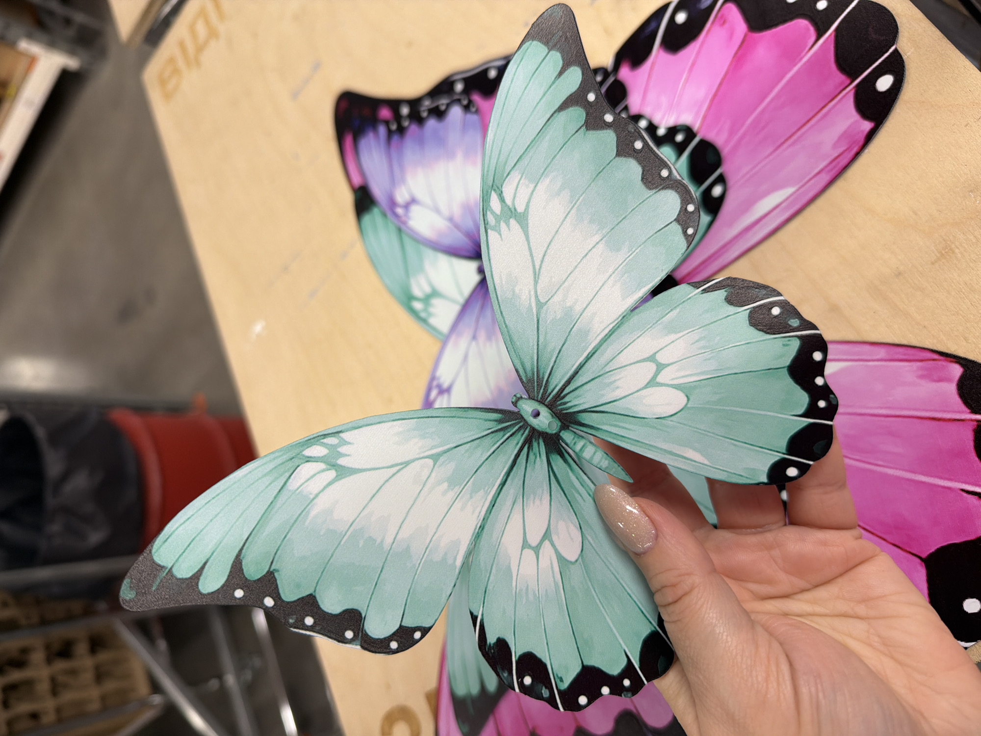 A close-up shot shows a hand holding the left wing of a large, teal and white butterfly decoration. The butterfly has black edges with white dots. Behind it, other butterfly decorations are visible, with wings colored pink and purple. The butterflies appear to be artificial decorations. They are placed on a light-colored wooden surface. In the blurred background, a dark trash can and red containers are visible, along with what appears to be shelving units. The scene does not provide any specific indicators of location in Ternopil, Ukraine.