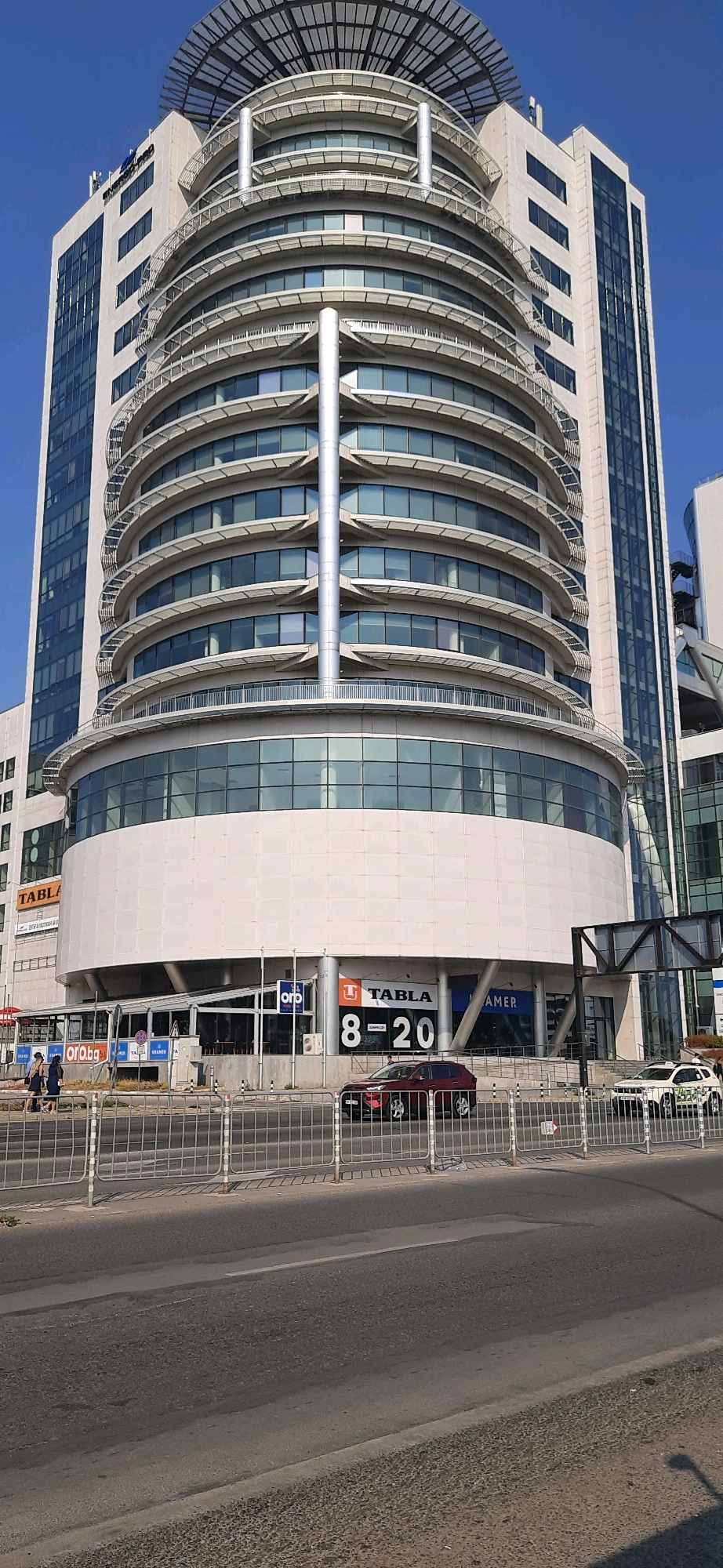 The image shows the "Grand Mall" in Varna, Bulgaria, from a street-level perspective. The mall is a large, modern building with a distinctive curved facade featuring multiple floors of windows and balconies. To the left of the curved section is a taller, rectangular glass-fronted building, which appears to be part of the same complex. A helipad structure is visible on the roof of the curved building. In front of the mall, at street level, is a storefront area with signage. Visible signs include "ORO.BG", "TABLA", and "AMER". The number "8-20" is prominently displayed with a "TABLA" logo.
On the street in front of the mall, a red SUV is parked in front of the "TABLA" store. A white vehicle, possibly a taxi, is parked further to the right. Several people are walking on the sidewalk in front of the mall, and a metal fence separates the sidewalk from the road. The sky is clear and blue, indicating a sunny day. The road in the foreground is asphalt with white lane markings.