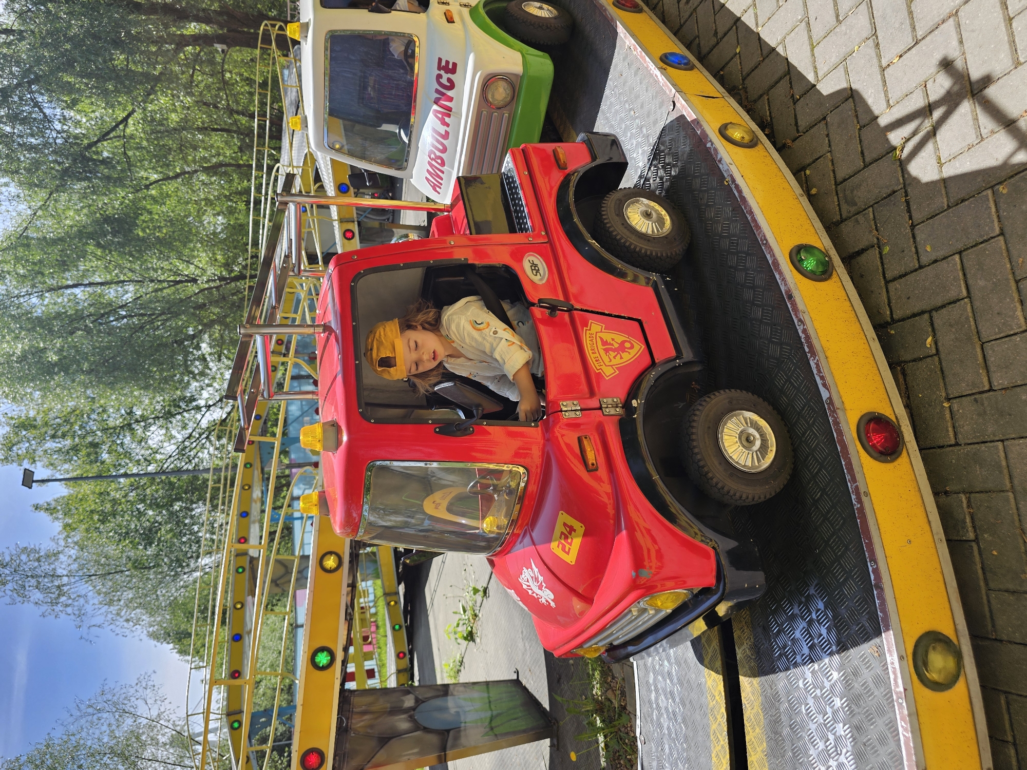 A child wearing a yellow cap is seated in a red fire truck on a ride at an amusement park or fair. Next to the fire truck is a white and green ambulance vehicle. The ride features a yellow border with colored lights. The ride is located outdoors, with trees visible in the background. The specific location is Khimki, Russia.