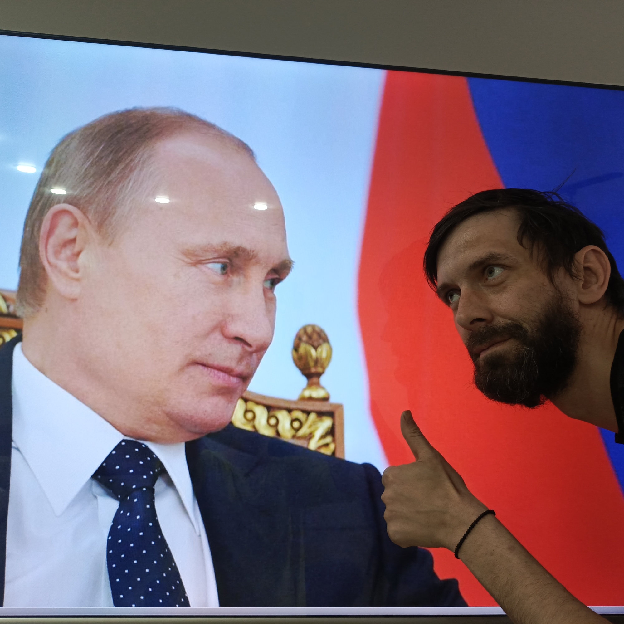 A color photograph shows a man standing next to a television screen displaying an image of Vladimir Putin. The man is fair-skinned with dark hair and a beard, and is giving a thumbs-up. He is wearing a black shirt and has a black bracelet on his right wrist. The television screen displays a portrait of Putin, who is wearing a dark suit, a white shirt, and a navy blue tie with white dots. The Russian flag is partially visible in the background behind Putin. The setting is indoors, likely in Марковский, Russia.