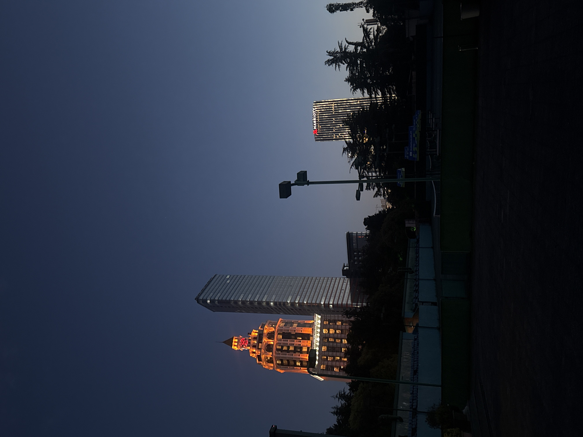 The image shows a nighttime cityscape of Batumi, Georgia. Prominently featured are two brightly lit buildings: the Radisson Blu Hotel, a tall, modern skyscraper with a grid-like facade, and the Sheraton Hotel, an ornate building with a unique, multi-tiered structure and a clock tower, illuminated in warm orange light. The "Marriott" sign is visible on another building. The sky is a gradient of dark blue. Streetlights are present along the street. Trees are around the hotel buildings, and a dark wall is visible in the foreground.