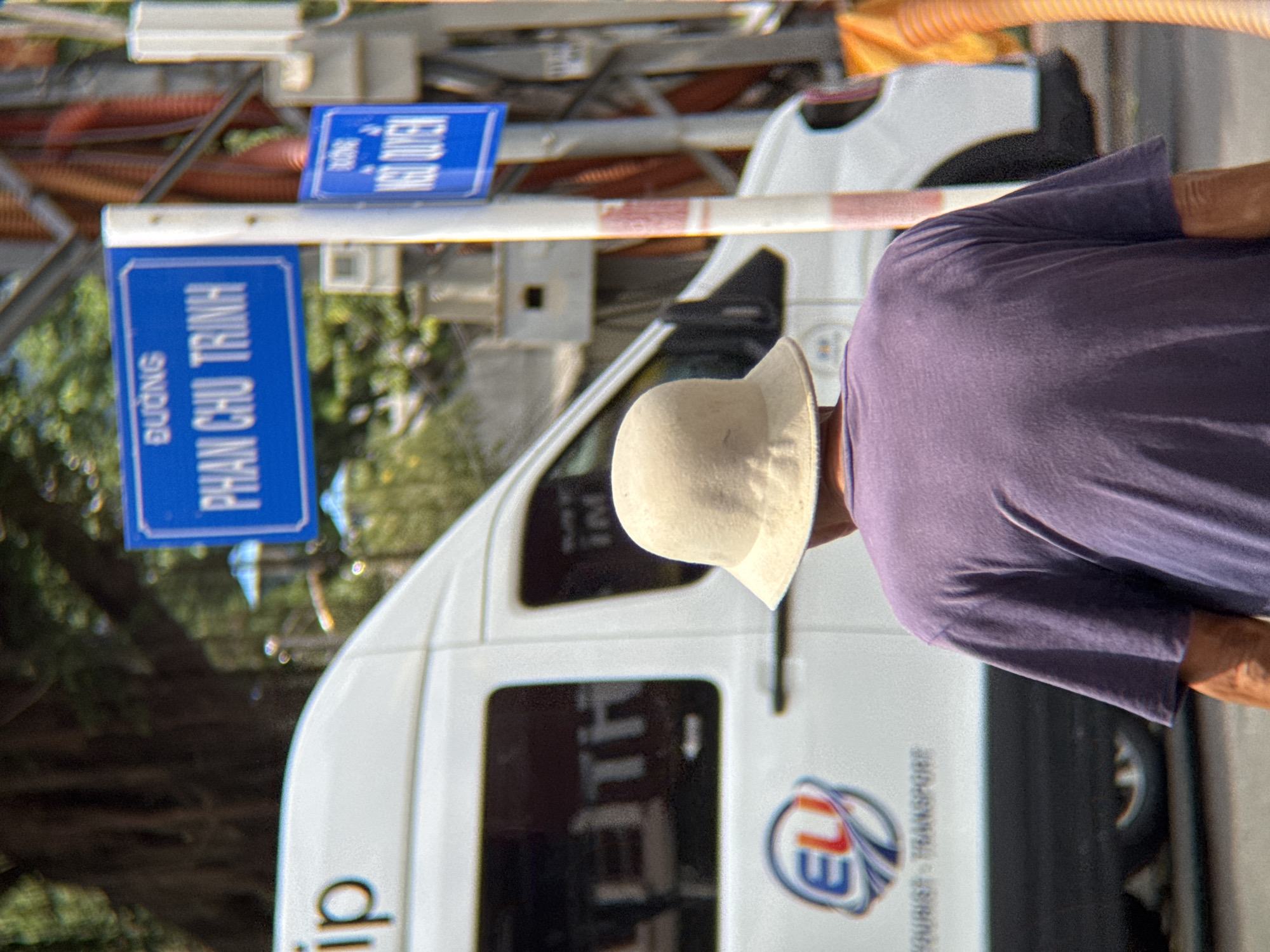 This is a medium-shot, eye-level outdoor photograph taken on a street in Nha Trang, Vietnam. The scene is dominated by a white van with tinted windows. On the side of the van, a circular logo with "EU" is visible, along with the words "TOURIST - TRANSPORT." The van appears to be parked or stopped.
In the foreground, from the back, a person wearing a purple t-shirt and a light-colored, wide-brimmed hat is visible. The person's head is tilted slightly down. Their arms are not clearly visible.
Behind the van and the person, two blue street signs are mounted on a white pole with red stripes. The upper sign reads "ĐƯỜNG PHAN CHU TRINH." The lower sign reads "ĐƯỜNG NGÔ QUYỀN." These are street names in Nha Trang.
The background is blurred, showing greenery, likely trees and foliage, suggesting a sunny day with dappled light. There are some vertical red elements visible to the right of the street signs, possibly part of a structure or utility pole. The ground appears to be asphalt. The overall impression is a street scene in a Vietnamese city.